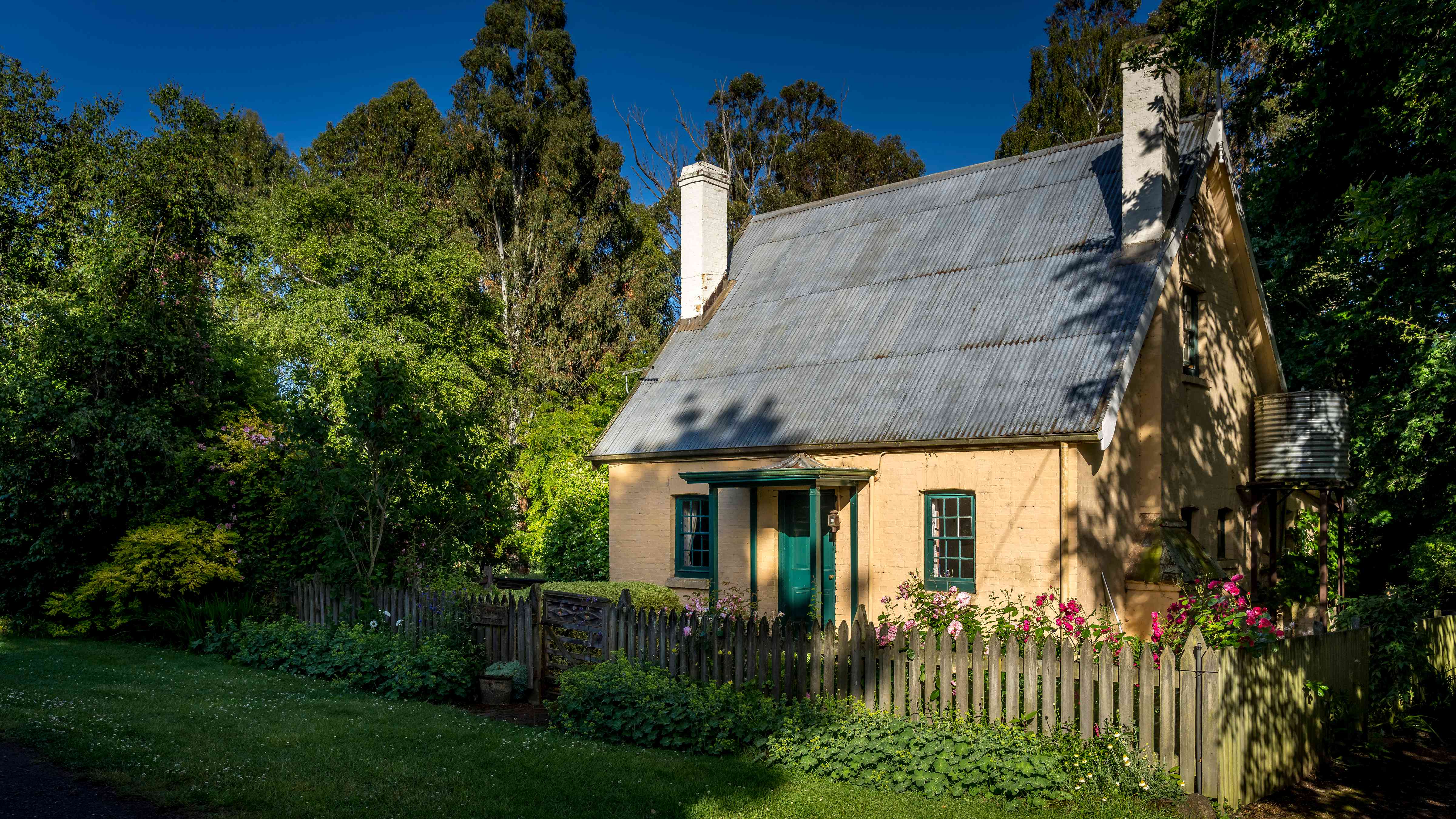 The cottage is surrounded by trees and a cottage style garden with pink roses. A timber picket fence runs along the front garden with alchamila plants and a lawn in the foreground. The steep pitched corrugated iron roof has two chimneys on either side of the house. The front of the cottage has two paned windows painted green with a green front door in the middle and a small porch over. The walls are limewashed brick and a tank stand is on the right of the cottage. Photo: Rob Burnett.