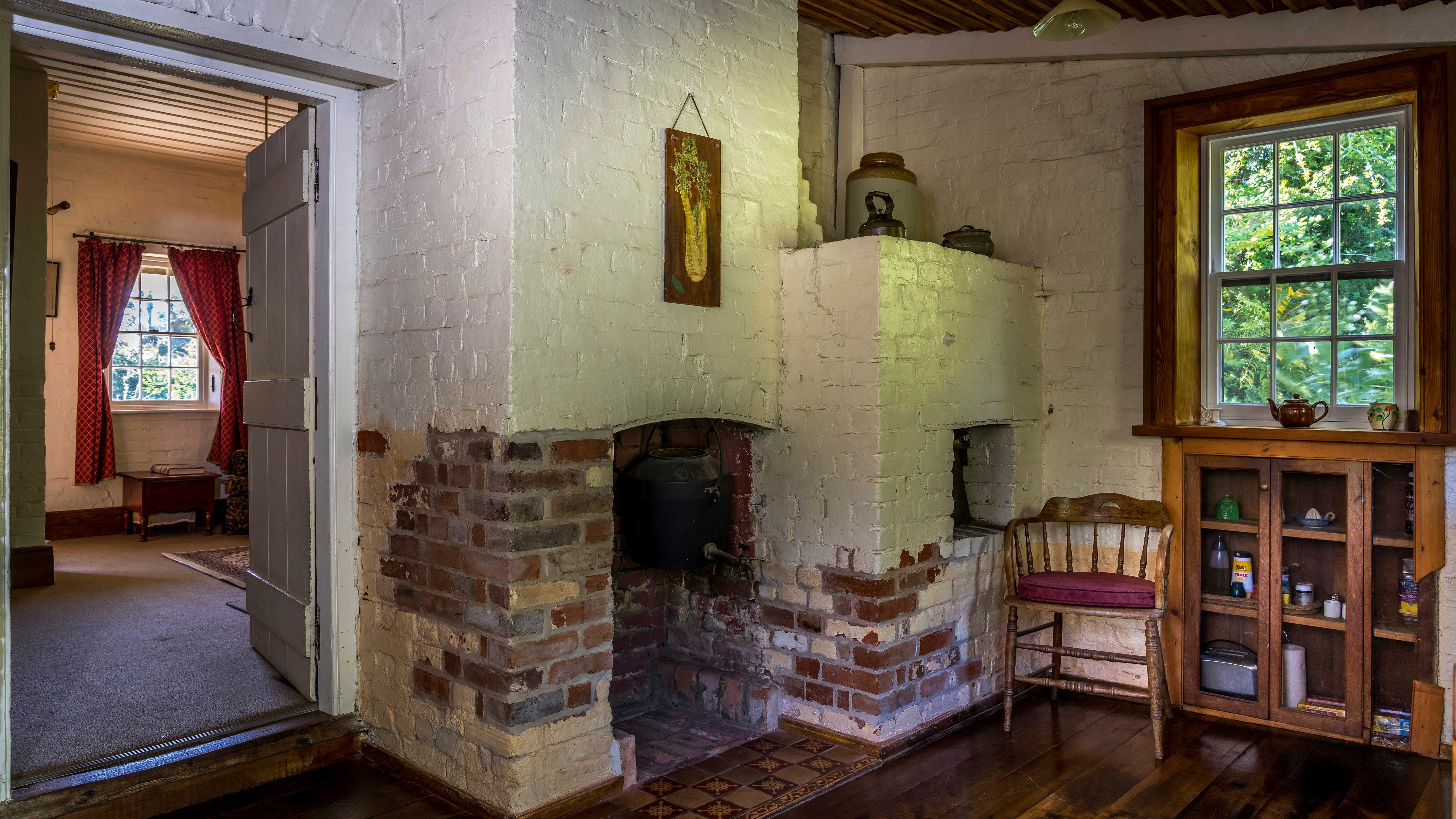A timber surrounded paned window has a safe style cupboard below with condiments. A wooden captain’s chair with a maroon cushion is to the side. Brick walls painted white and unpainted have a fireplace and a bread oven with several kitchen items sitting on top. A picture of a bunch of celery hangs on the wall. On the left of the image is a doorway leading to the sitting room which has brown carpet and a window with red curtains pulled open. Photo: Rob Burnett.