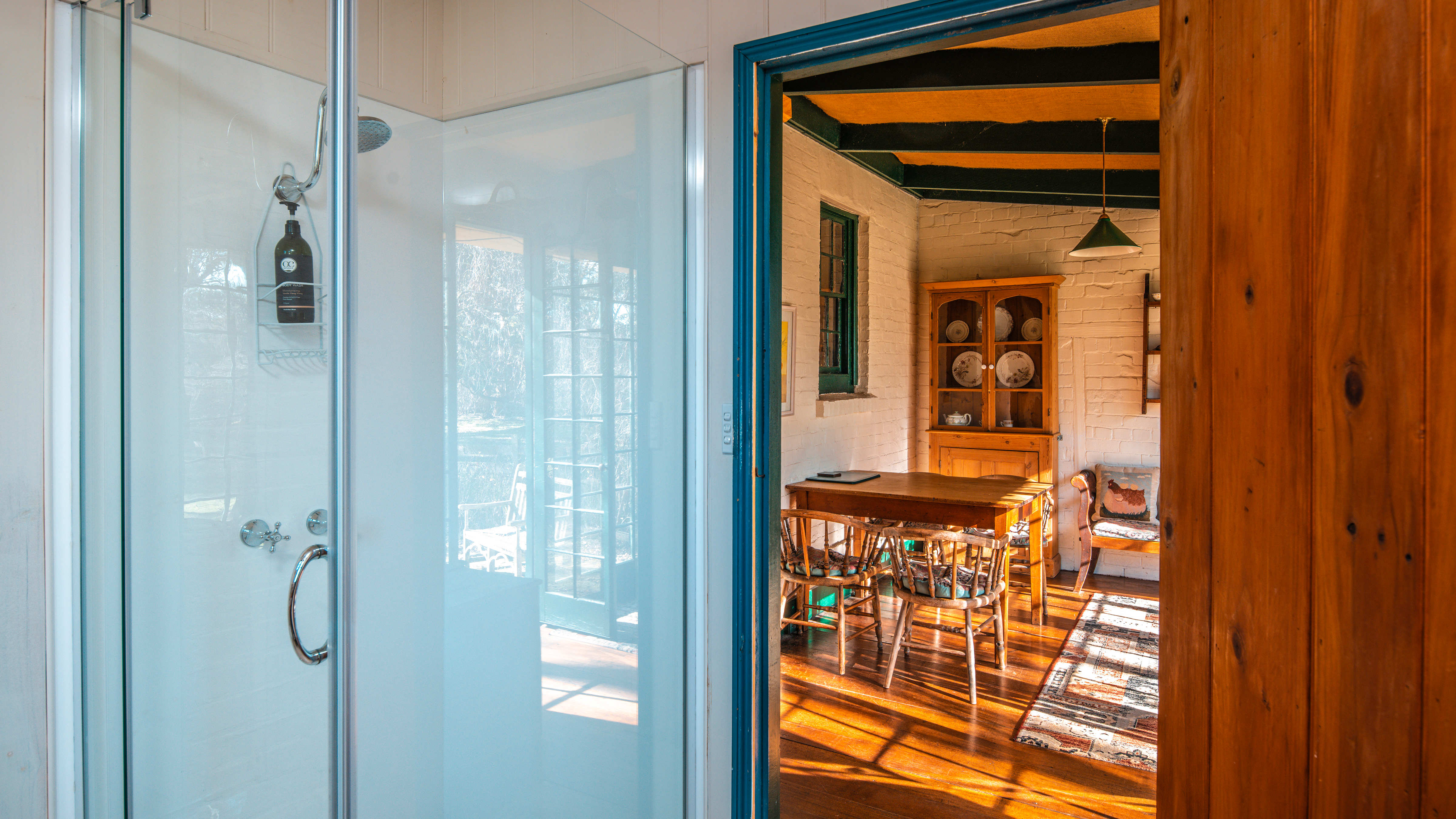 To the left of the image is a glass shower screen with a view through the timber bathroom door to the sunroom. A timber table with antique chairs around is in front of a pine dresser with antique plates displayed. A rug is on the floor boards. Photo: Rob Burnett.