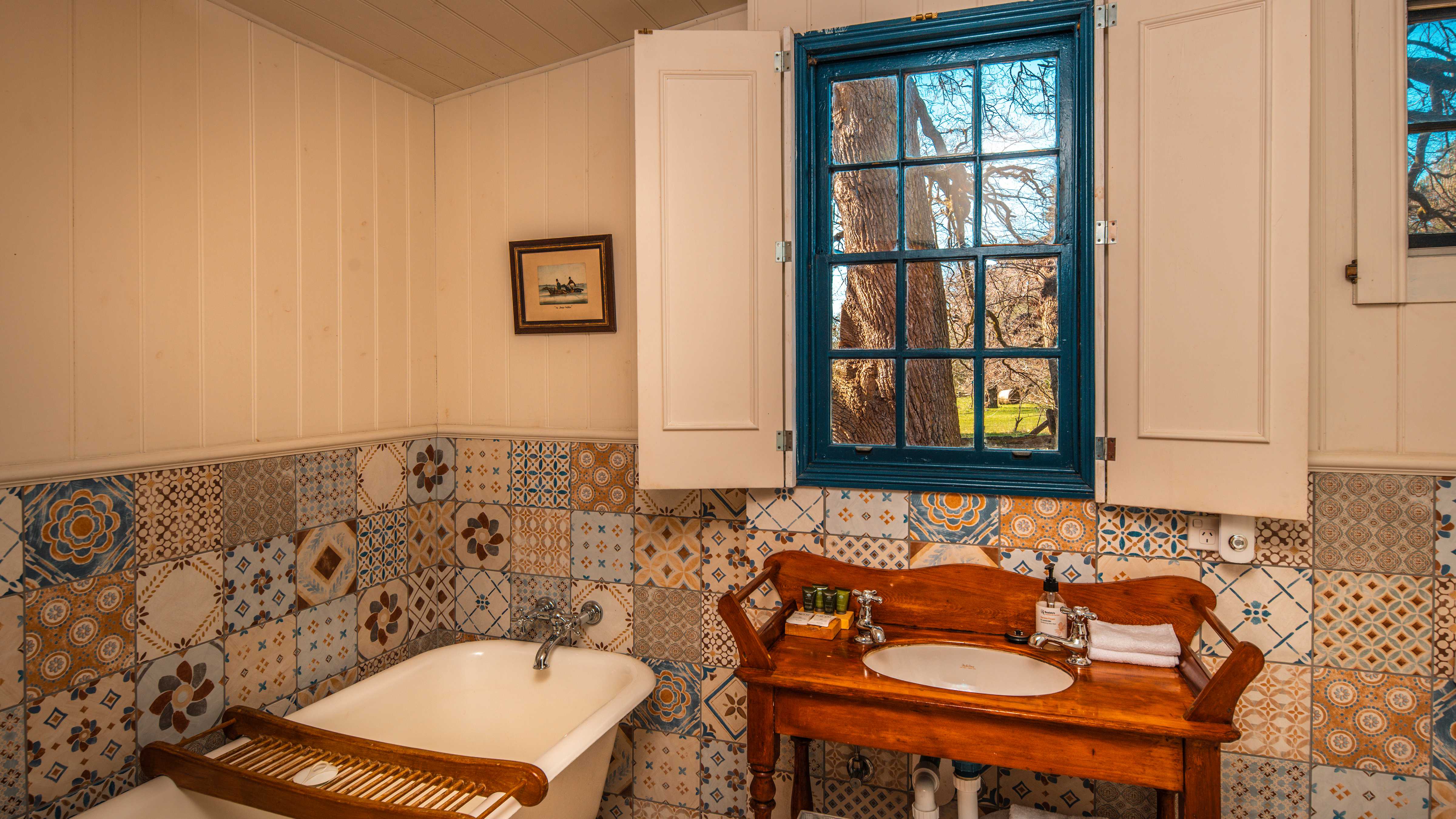 The bathroom walls have a decorative tiling dedo with white painted timber panelling above. The blue painted paned window has shutters on the side and looks out to a large tree and field beyond. A timber washstand has a white oval basin with two pillar taps and a selection of bathroom items on top. An old fashioned white bath has a timber rack across it. Photo: Rob Burnett.