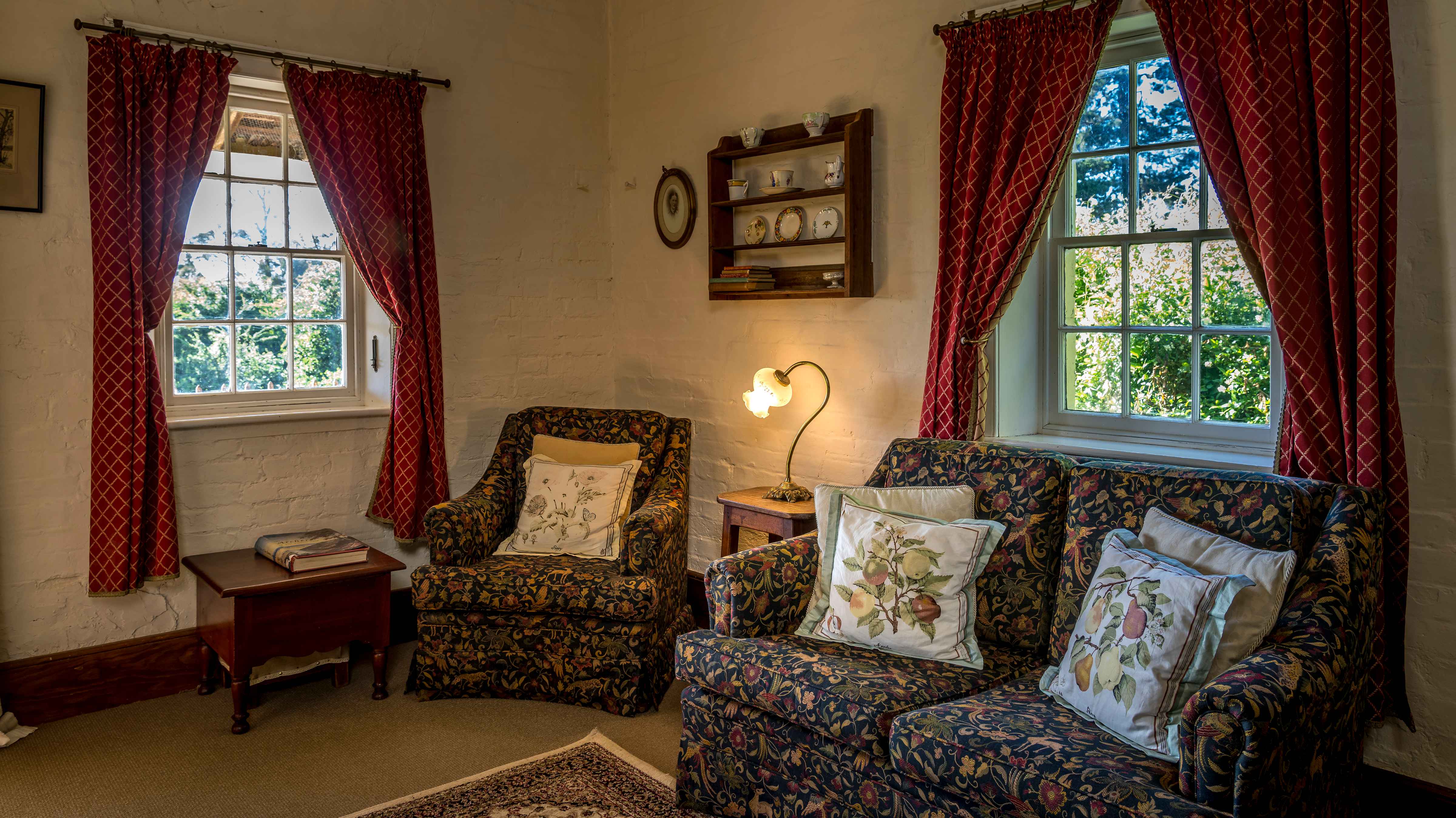 This room is decorated in the cottage style with a small dark pattern fabric covered couch and single chair. Cushions with pear tree motifs are on the couch and chair. A lamp which is turned on is lighting the wall which has a small set of timber shelves decorated with old fashioned cups and saucers and a couple of little books. Two Georgian paned windows have red curtains pulled to each side. Green foliage can be seen through the windows. A timber box with a large book stands beside the chair and under the window. Photo: Rob Burnett.