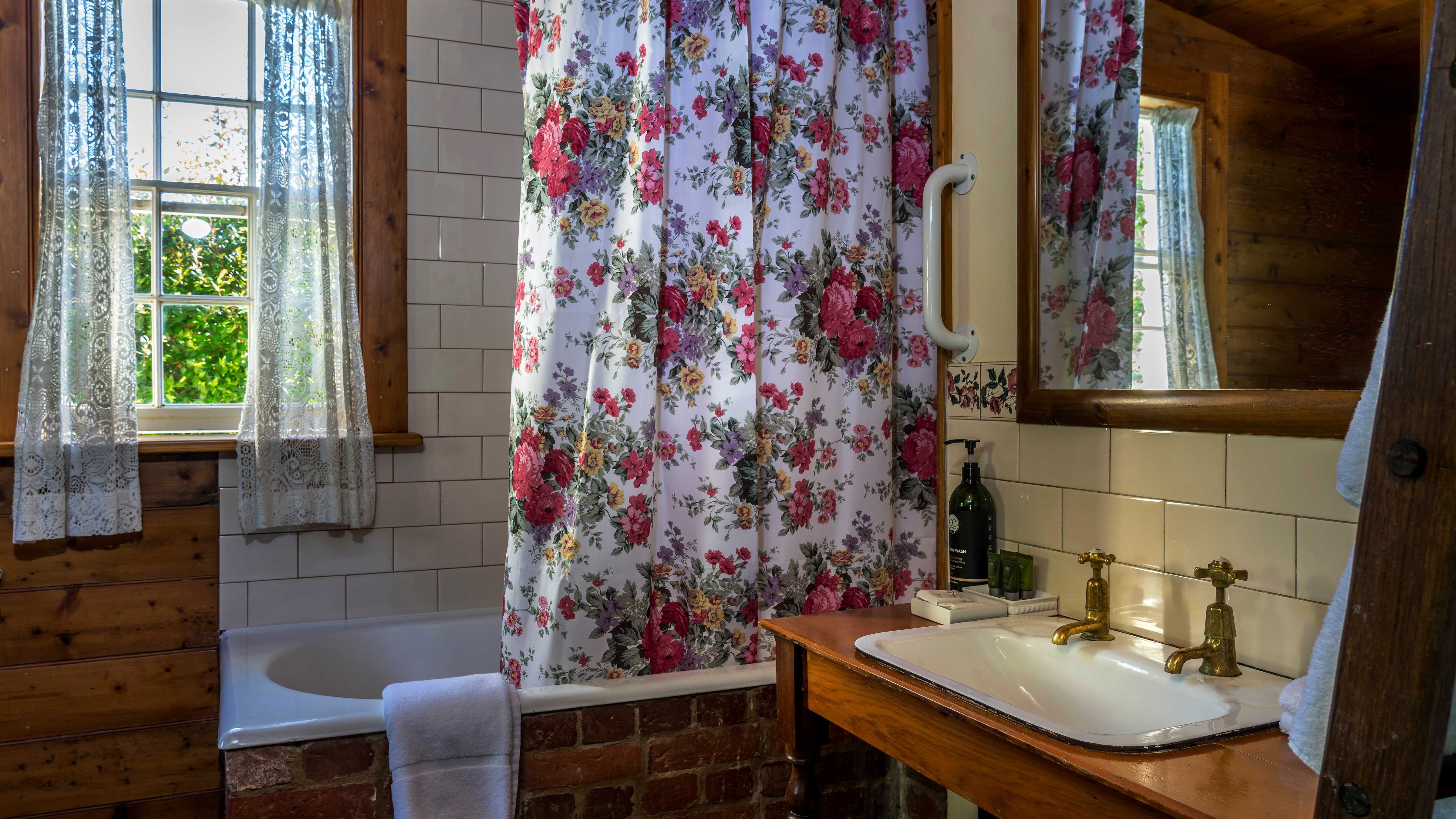 An old fashioned bath is enclosed with bricks and a floral shower curtain hangs in front of a white tiled wall. A Georgian paned window has lace curtains hanging with a view to green foliage. On the left is a timber washstand with a white basin and two pillar taps with a mirror above. Photo: Rob Burnett.