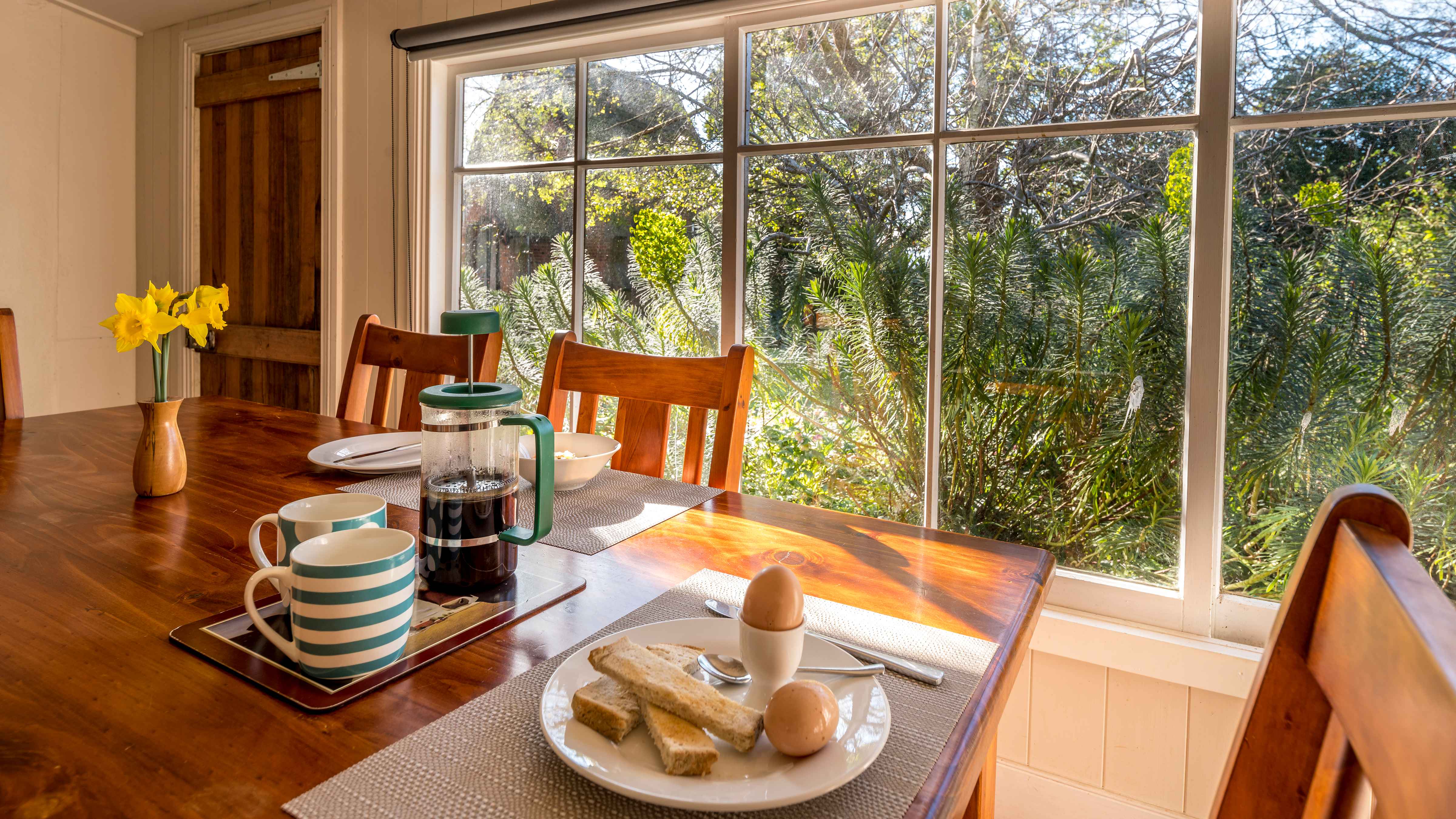 A timber table is set for breakfast with blue and white mugs, coffee plunger half full of coffee,two boiled eggs, one in an egg cup and toast on a white plate. There are three chairs around the table and a small vase of daffodils is on the table. The large paned windows are looking out to the garden which has pale green euphorbias in bloom. A timber door which opens to the garden is closed. Photo: Rob Burnett.
