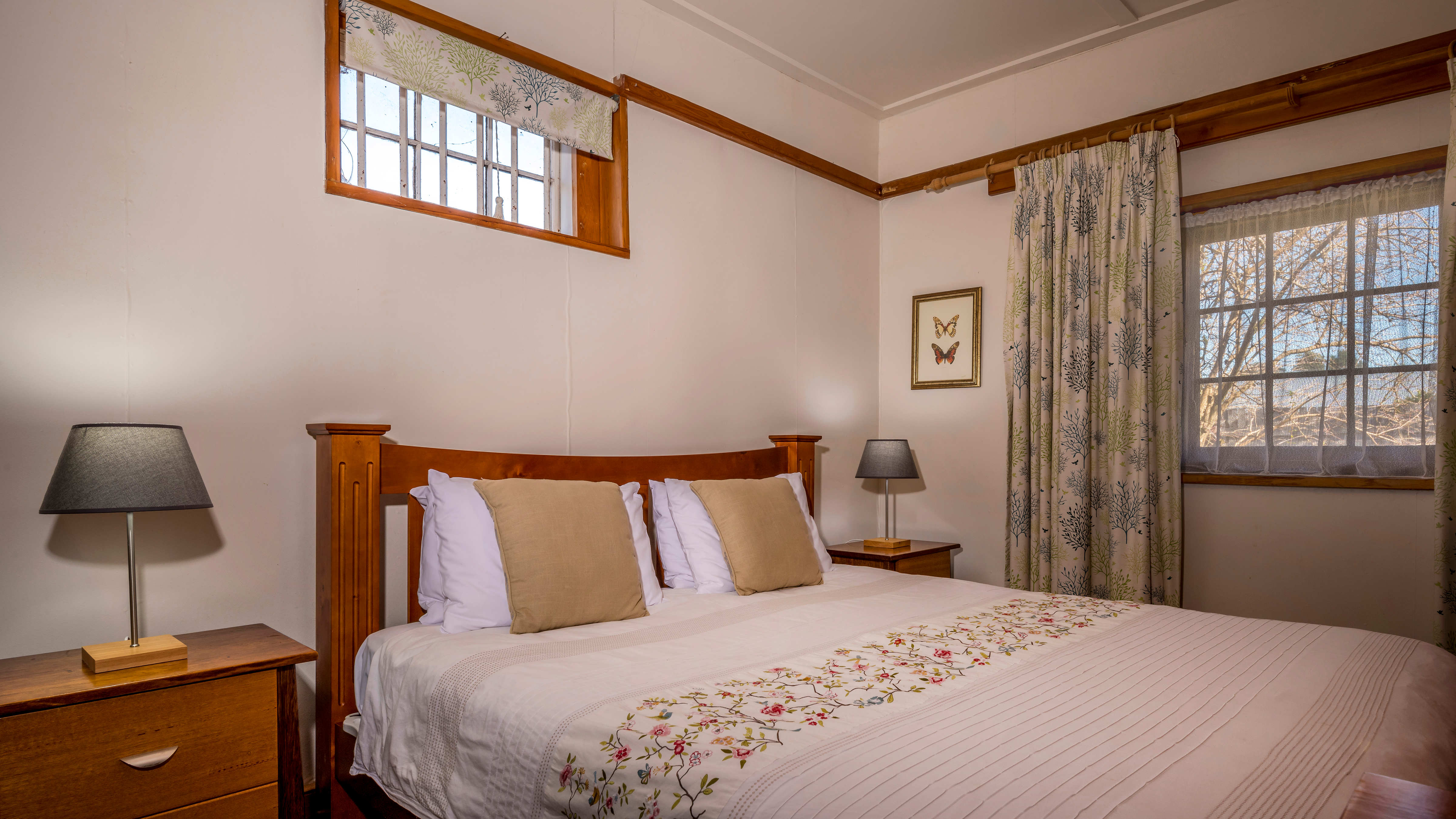 A king size timber bed stands in the middle of the image which has a white and floral patterned quilt cover and two fawn coloured cushions. Two bedside drawer stands have two bedside lamps with grey shades. A small paned window with bars and a blind is above the bedhead and green and blue floral curtains hang beside a second larger window on the far side of the bed. A timber picture rail runs around the room and a butterfly print hangs on the wall. Photo: Rob Burnett.
