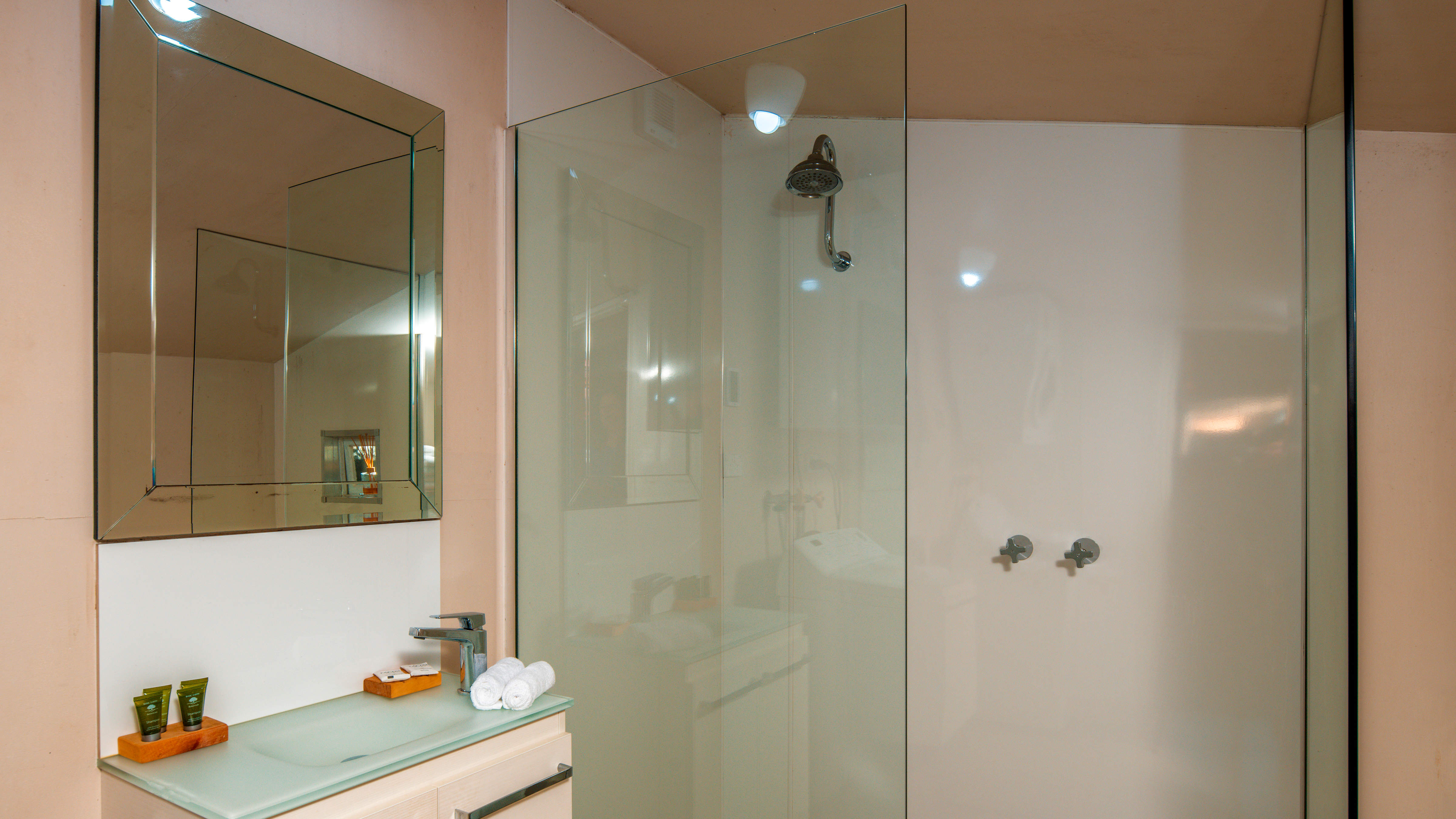 A large square mirror hangs on the wall above the basin. There are shampoo and conditioner bottles, soaps and face cloths on the basin alongside a chrome pillar tap. A clear glass shower screen has a wall mounted shower head and wall taps behind it. Photo: Rob Burnett.