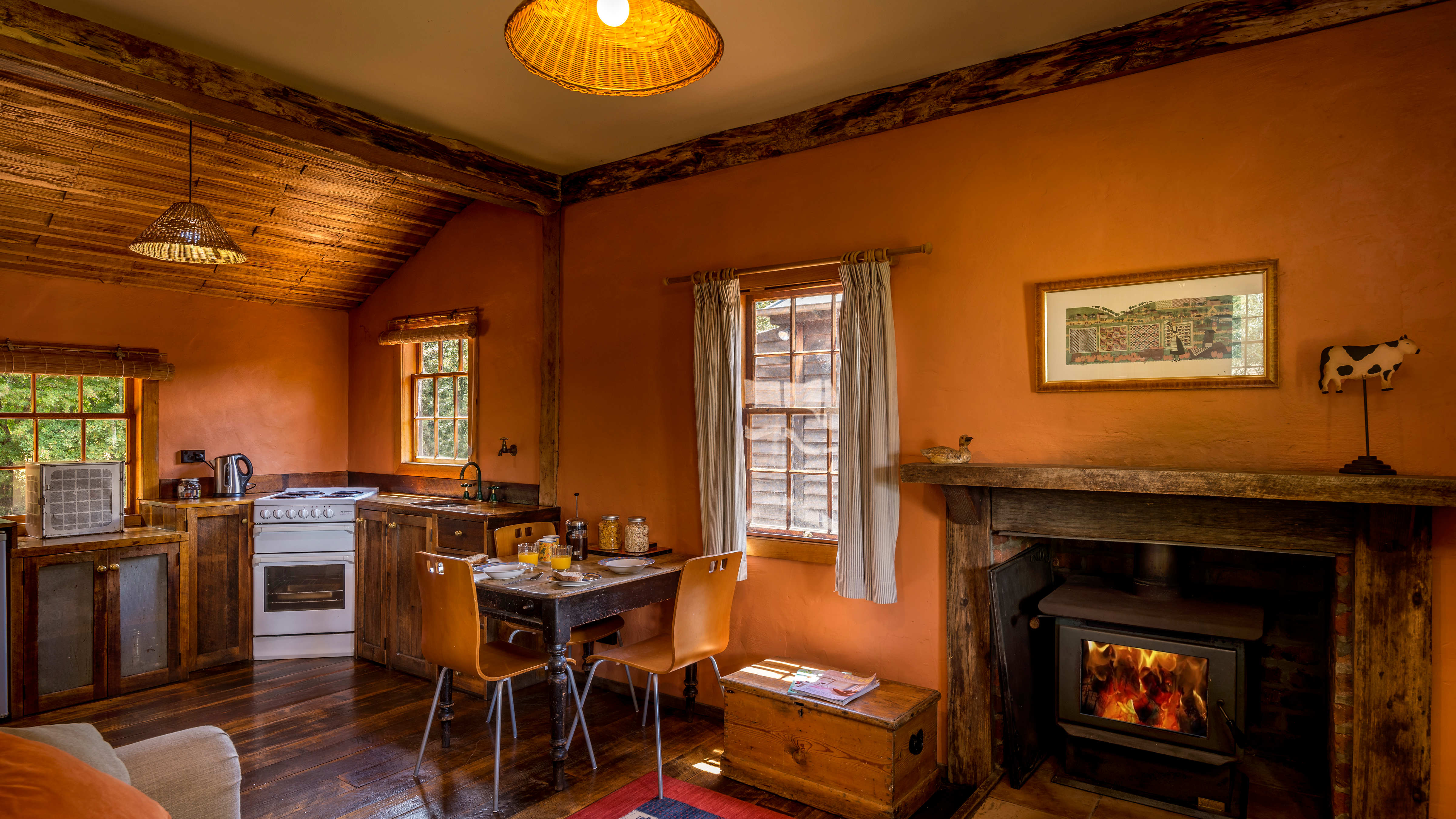 The open plan farmhouse kitchen has timber cupboards and table and chairs are bordered by georgian paned windows and terracotta painted walls. The table is laid with breakfast provisions including jars of cereal, glasses of orange juice, bowls and plates with toast and a coffee plunger. To the right of the image is the wood heater which is alight with orange flames. The rough timber fireplace surround has a picture hanging above and decorator items on the mantle. A pine timber box sits between the fire and the kitchen dining table. Photo: Rob Burnett.