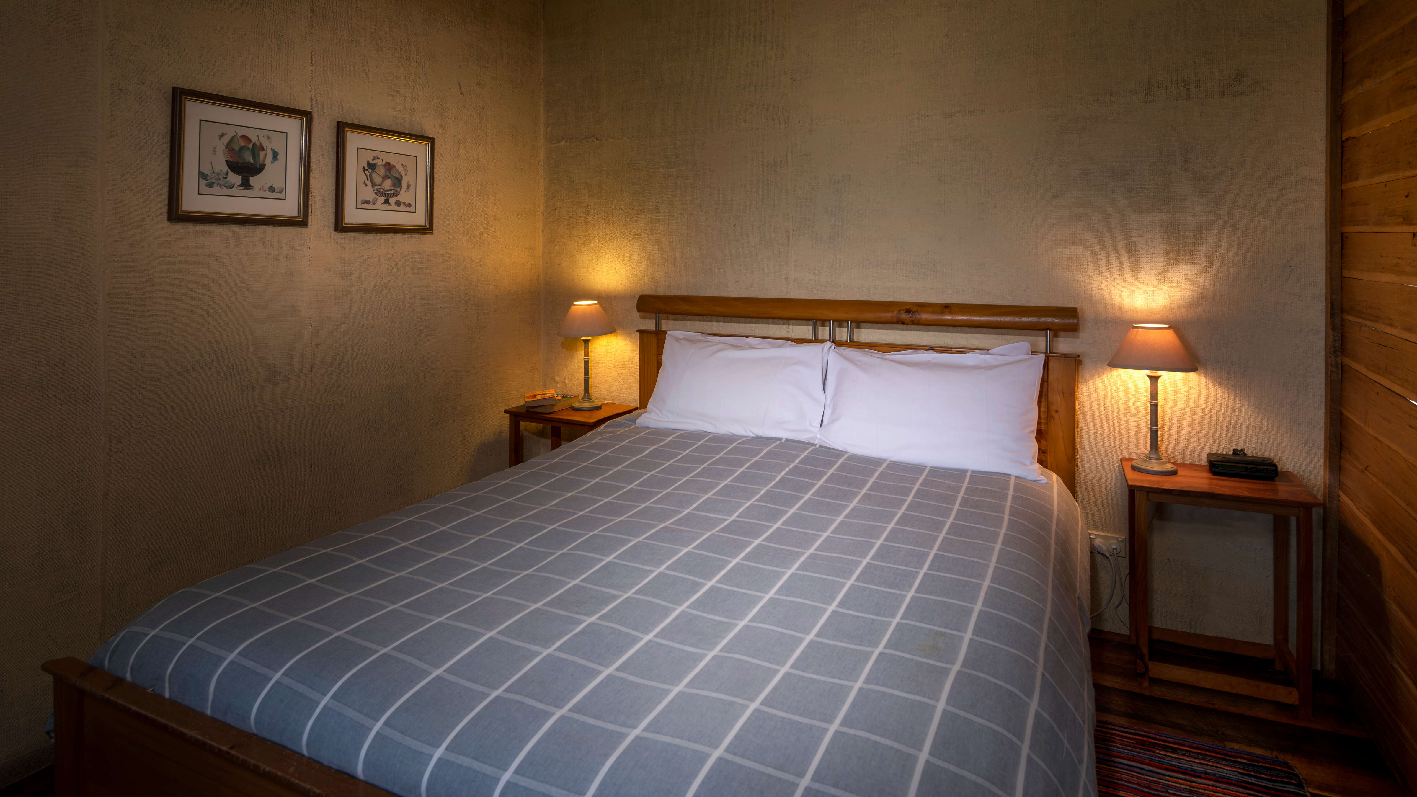 The bedroom walls are covered with painted hessian with two pictures hanging on the wall. The wall to the right hand side is timber horizontal palings. The timber framed bed in the centre has a blue and white checked quilt with white pillows. Timber bedside tables have bedside lights which are turned on. Photo: Rob Burnett.