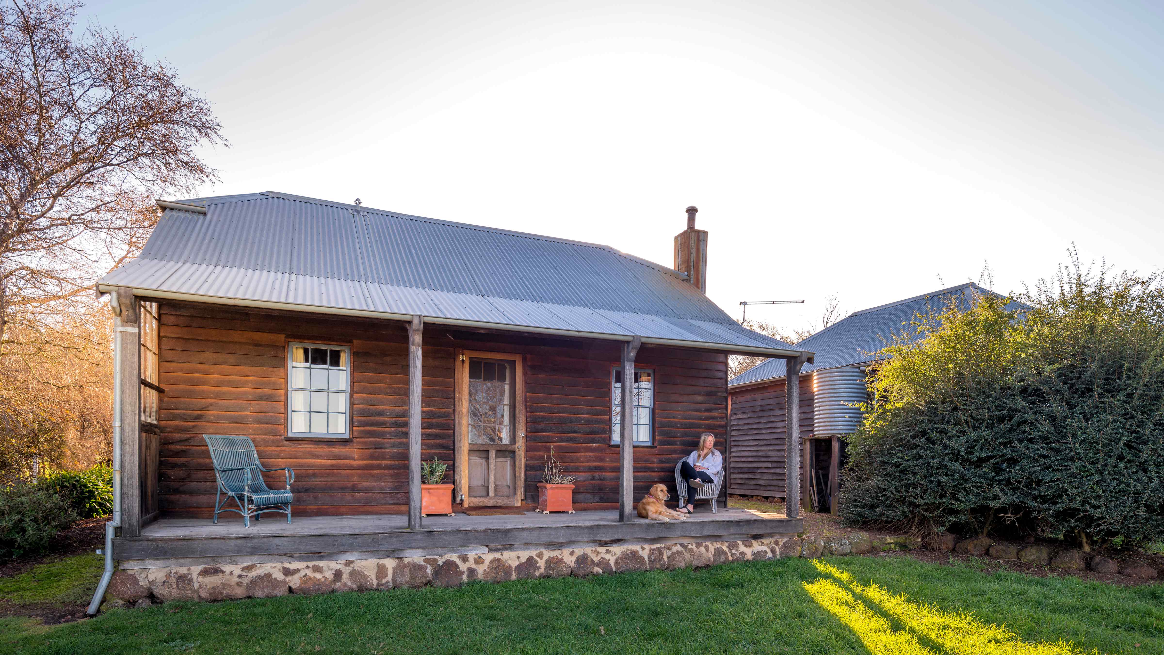 The external verandah of Pumpkin cottage has timber posts and horizontal weatherboard walls. Two windows and a timber front door with two square terracotta pots on either side A chimney stands to the right of the corrugated iron roof. A lady is sitting in a chair with a golden retriever; they are looking into the distance. A shaft of sunlight is shining through a gap beside the cottage on to the grass. Photo: Rob Burnett.