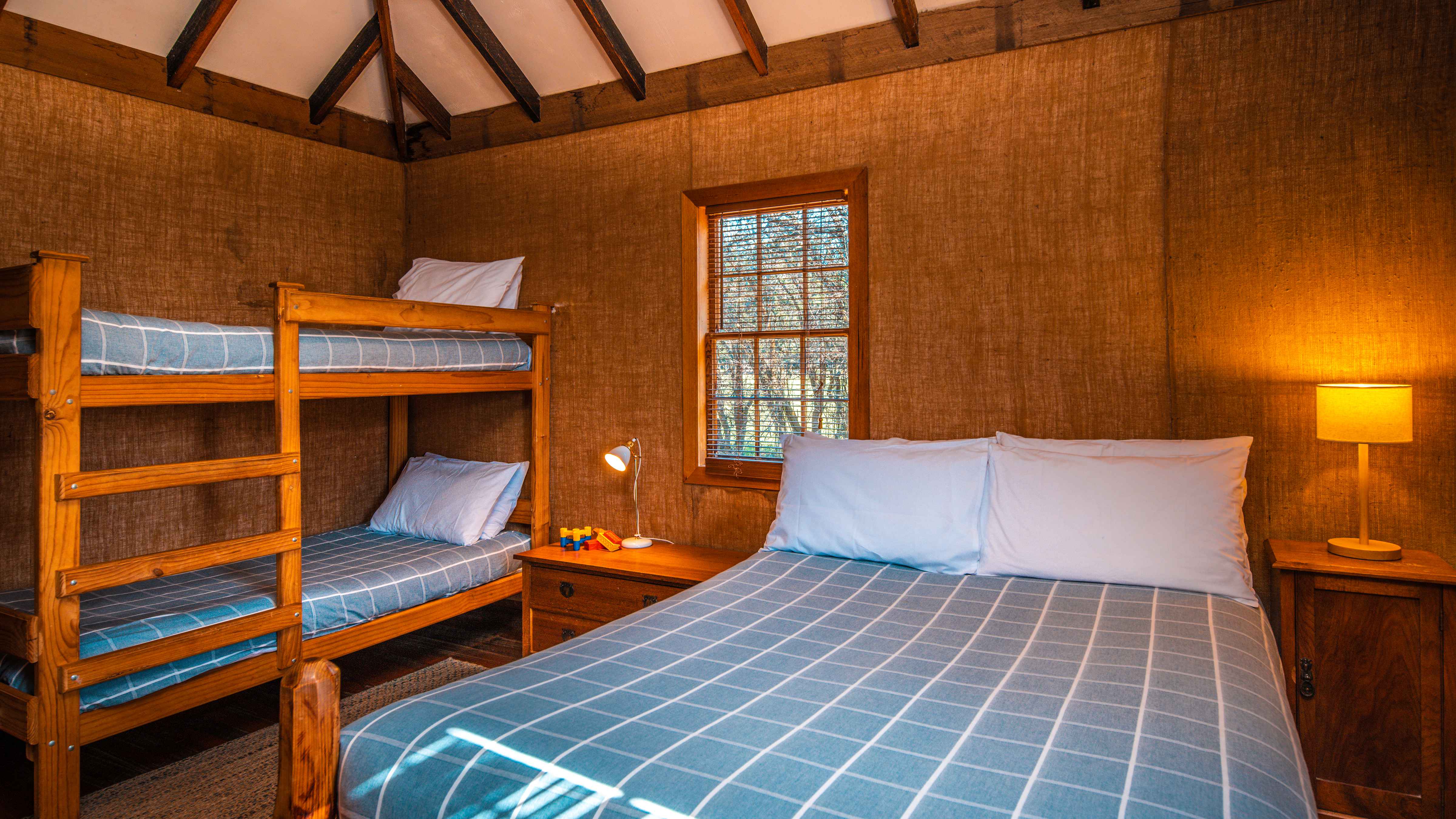 A double bed is in the foreground with a blue and white checked quilt cover and white pillows. To the left are a set of timber bunks with matching blue and white checked quilts and white pillows. The walls are covered in brown hessian and a window looks out to trees. A set of drawers with a lamp sits between the bed and the bunks and another bedside cupboard with a light sits to the right of the double bed. The ceiling is white with timber beams. Photo: Rob Burnett.