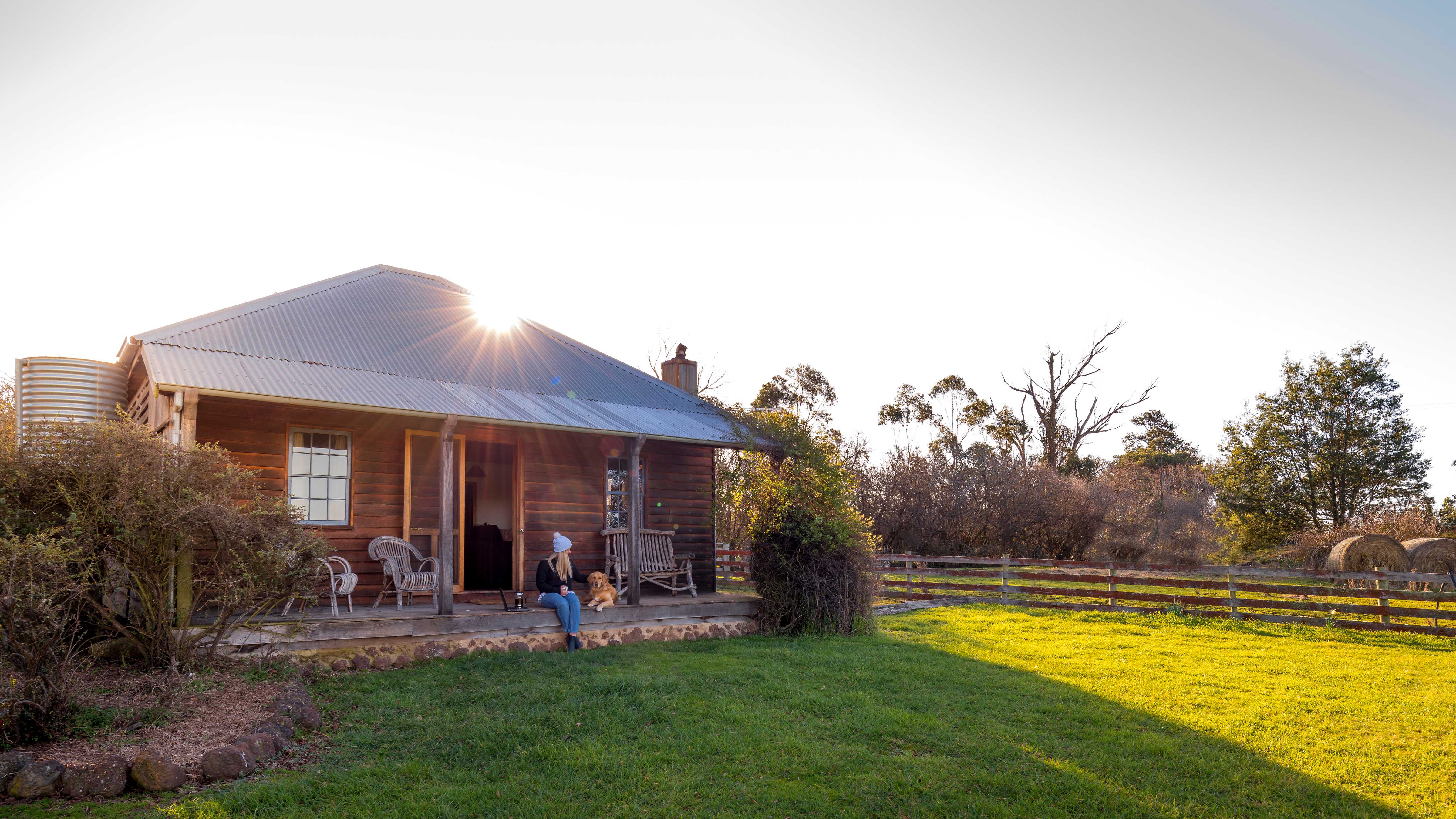 Lady and dog sitting on the verandah of the cottage. Sun is disappearing behind the corrugated iron roof. Timber walls have two windows and there are shrubs on either side of the verandah. A grassed area is in front and a timber fence is alongside the cottage with trees in the background. Photo: Rob Burnett.