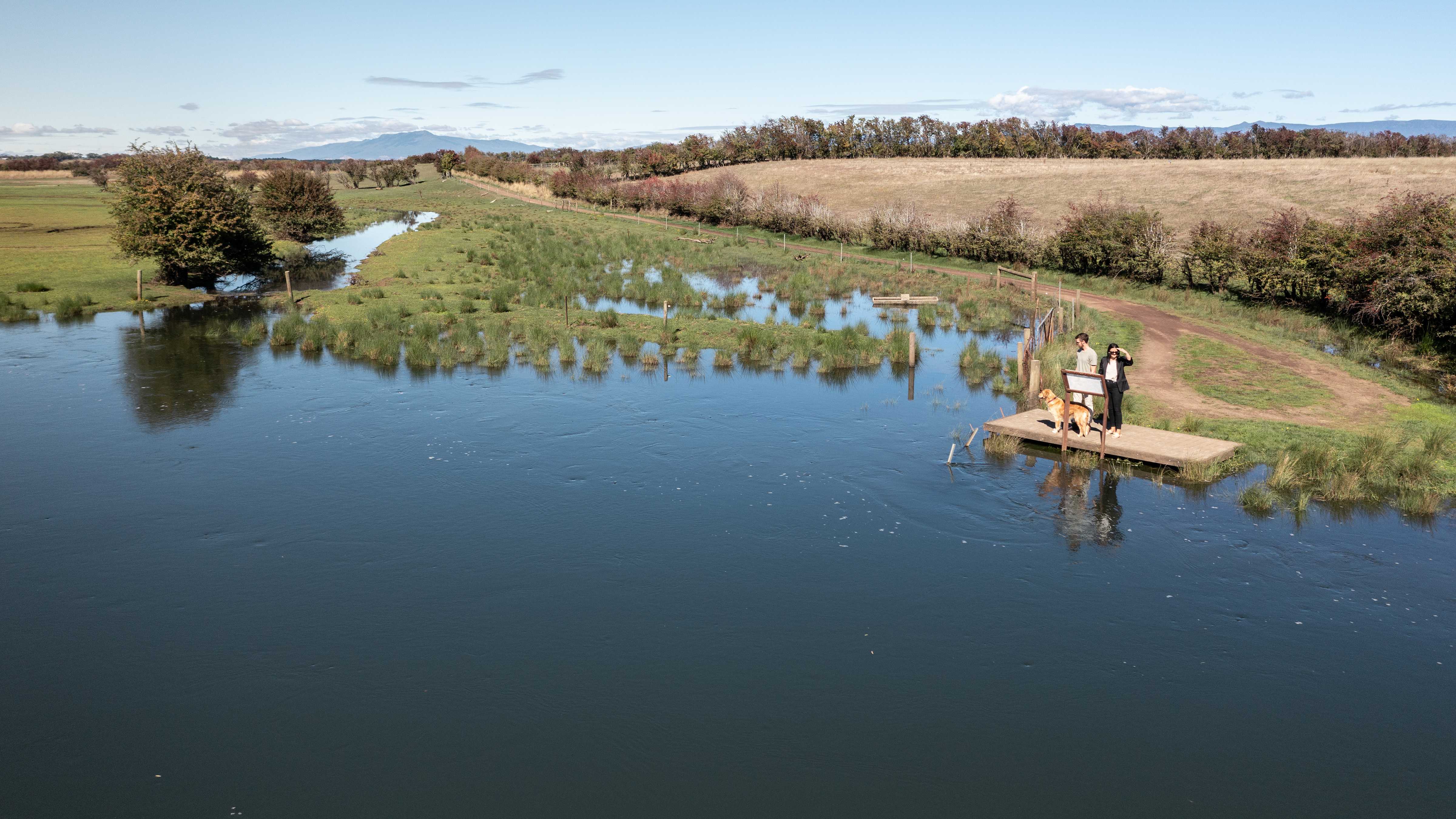 A couple and a dog stand on the jetty on the banks of the Macquarie River. The river is in low flood and creeps into the paddock alongside. Hedgerow lined paddocks continue in the distance. Photo: Kate von Stieglitz / Tourism Australia.