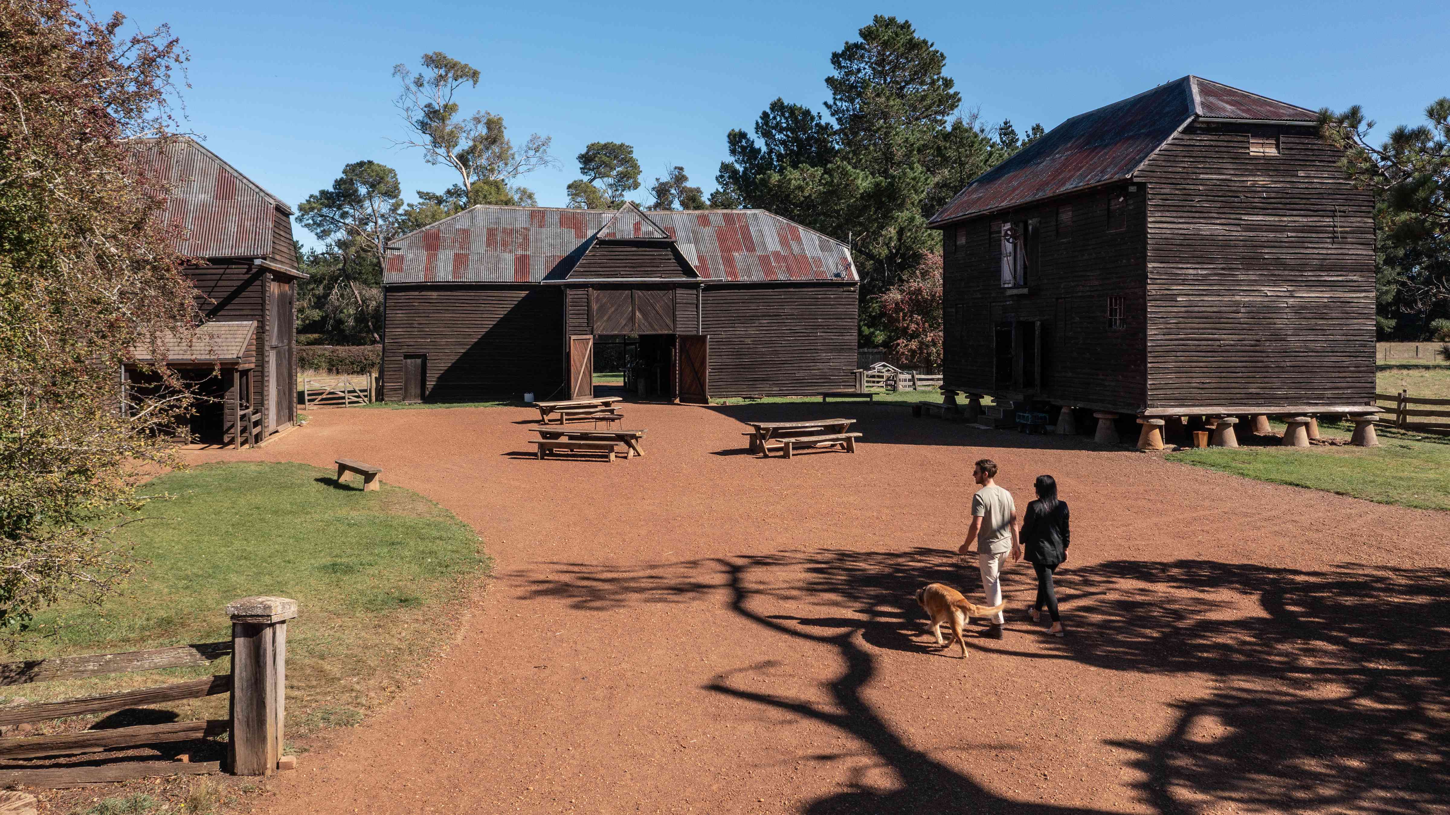 Two people and a dog are wandering into the quadrangle which is surrounded by the Sussex and Village barns and the Pillar granary.  The quad is gravel with 3 timber picnic tables in the centre. Photo: Kate von Stieglitz / Tourism Australia.
