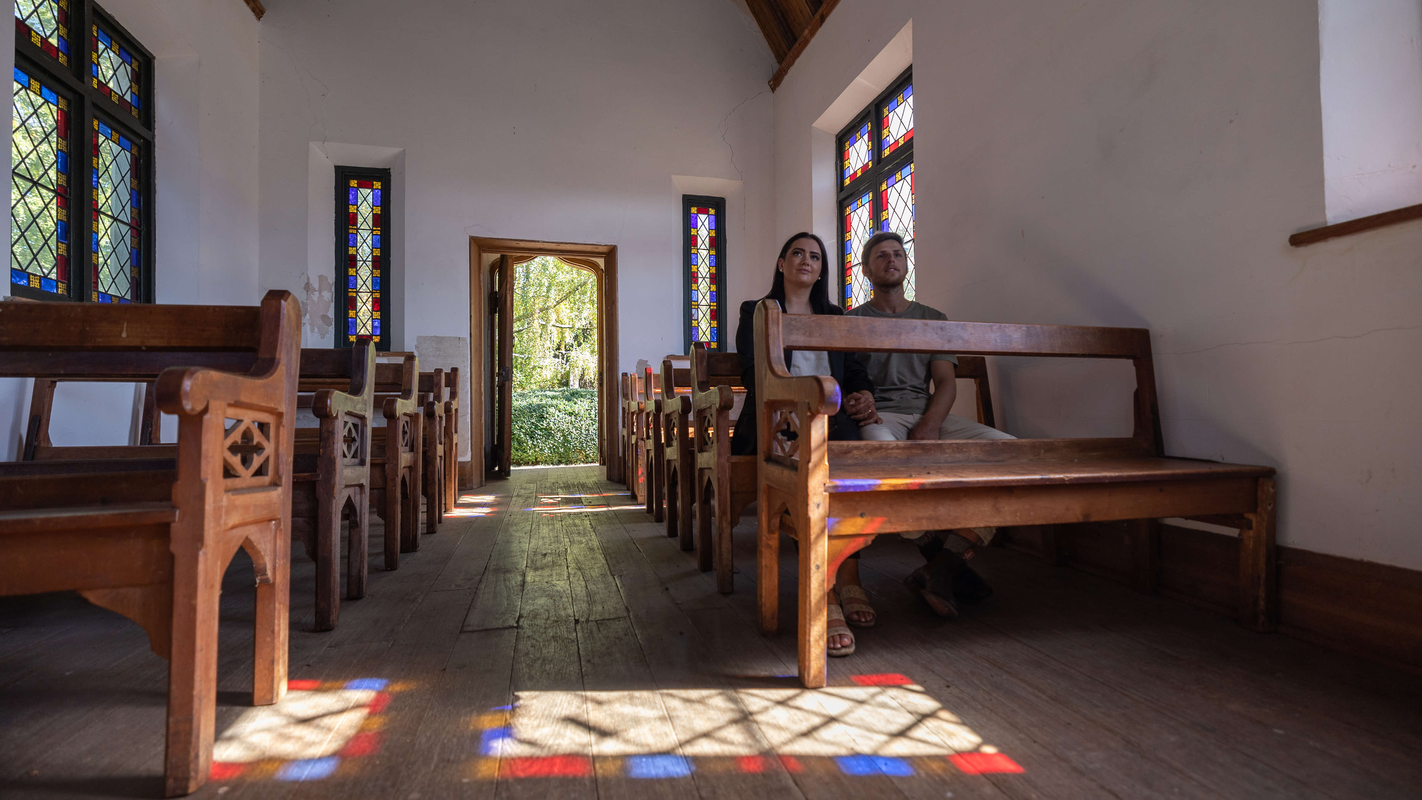 A couple sit on one of the huon pine pews in the Chapel circa 1850. The internal features include timber floor boards, white painted walls etched with stone blocks and a pine lined pitched ceiling.  The stained glass windows include blue, red and amber borders with diamond lead lighting. The sun coming through the windows cast coloured outlines on the floor. Photo: Kate von Stieglitz / Tourism Australia.