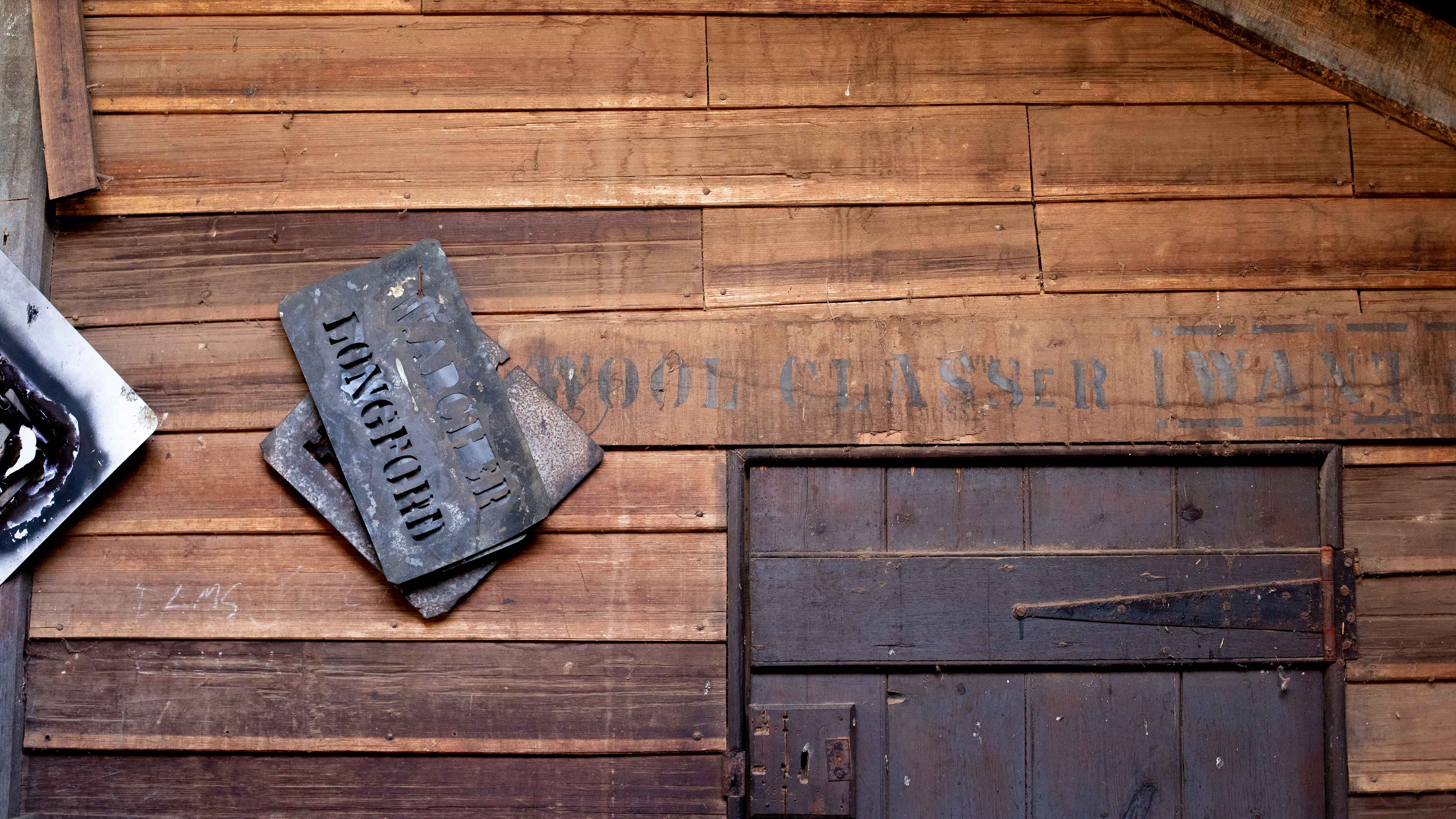 Metal wool brands with the wording ‘LONGFORD’ hang from a nail on a horizontal timber wall. A timber door with old fashioned hinges is also in the picture. Some printed words saying ‘WOOL CLASSER WA’ are on the wall in black paint. Photo: Kieran Bradley.