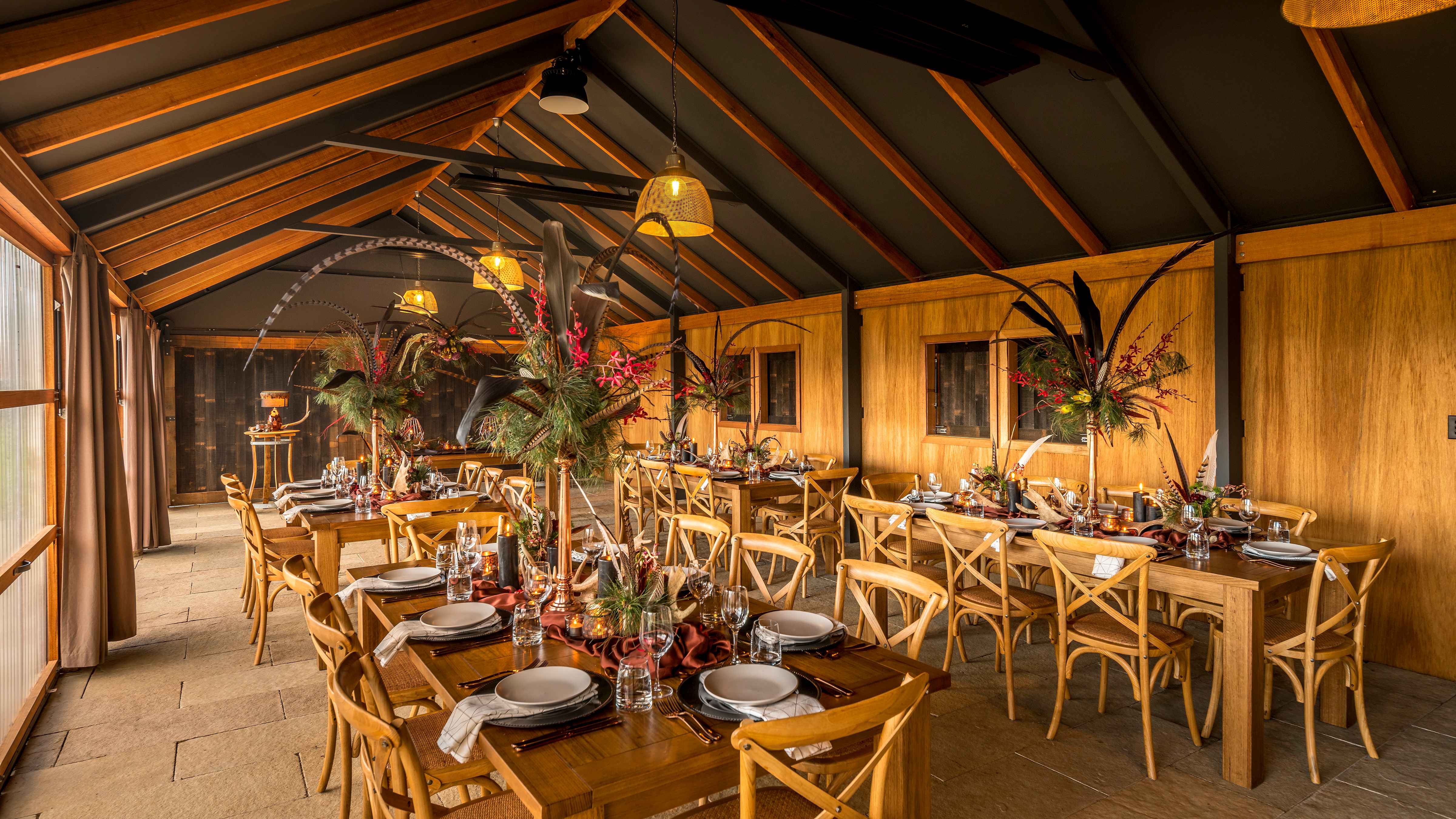Long wooden tables are set in The Archery with crockery, cutlery, glassware and large floral arrangements that also contain feathers. Wooden cross back chairs are placed at the tables and pendant lights hang from the ceiling. The floor is limestone pavers and exposed rafters line the ceiling. Photo: Rob Burnett.