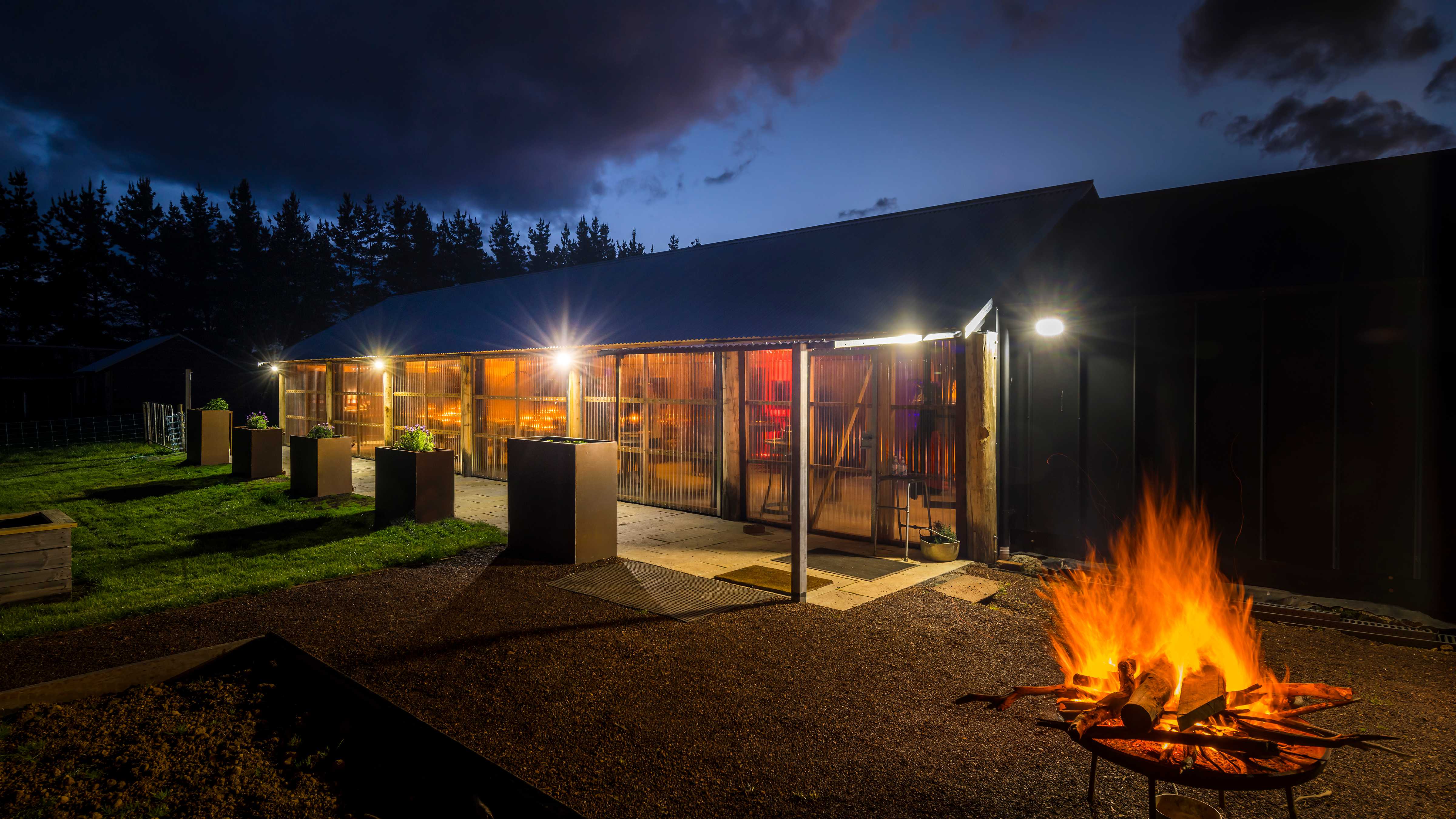 An exterior night time view of the Archery with a lit fire pot in front. The side of the building is clear laserlight and the lights are on inside. There is grass and gravel in front of the building and the darkened sky behind. Photo: Rob Burnett.