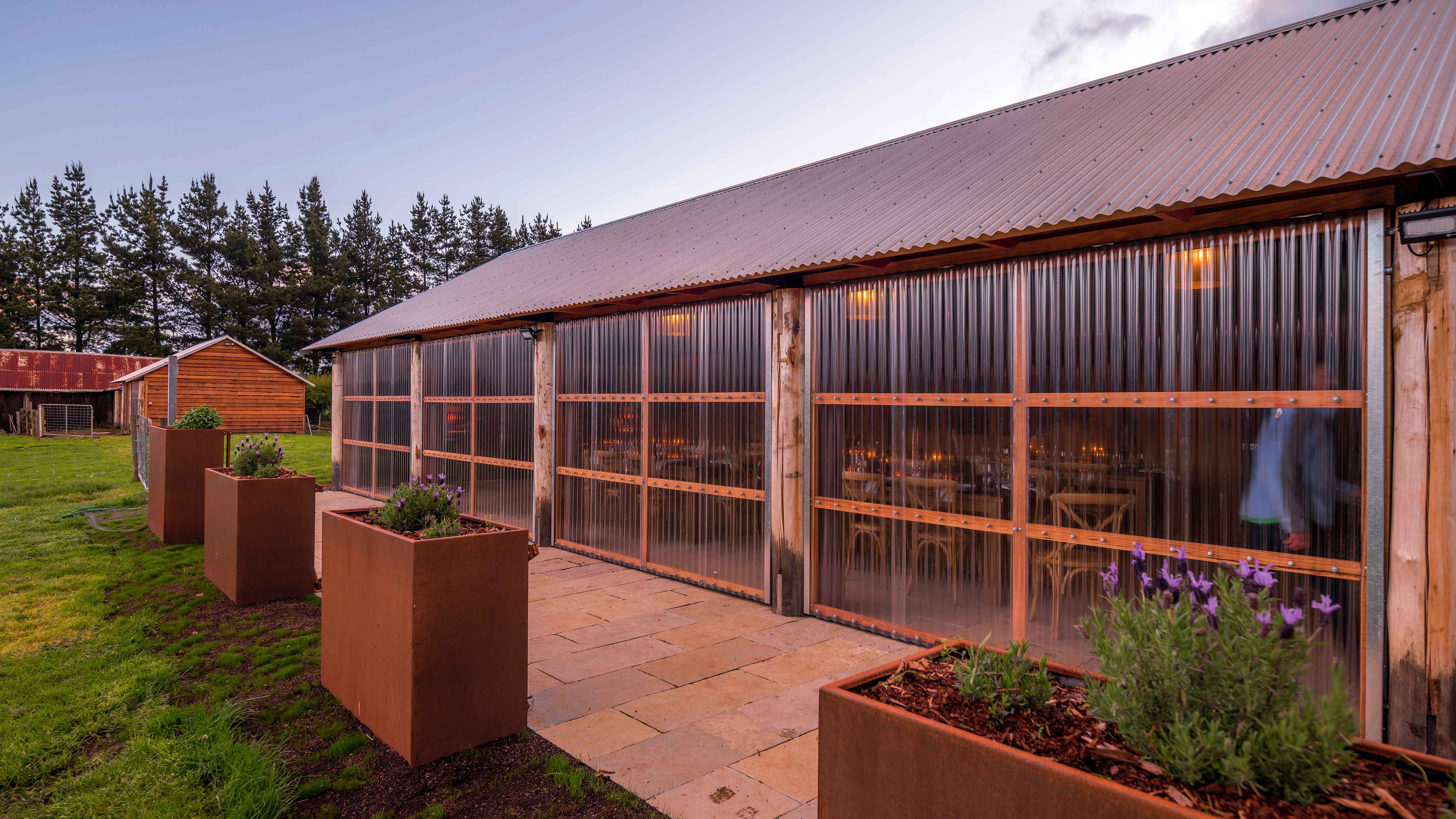 The Archery with its clear laserlight doors closed in the evening light. The building has a corrugated iron roof. Vertical corrugation laserlight walls and limestone pavers are bordered by iron planter boxes and grass. Photo: Rob Burnett.