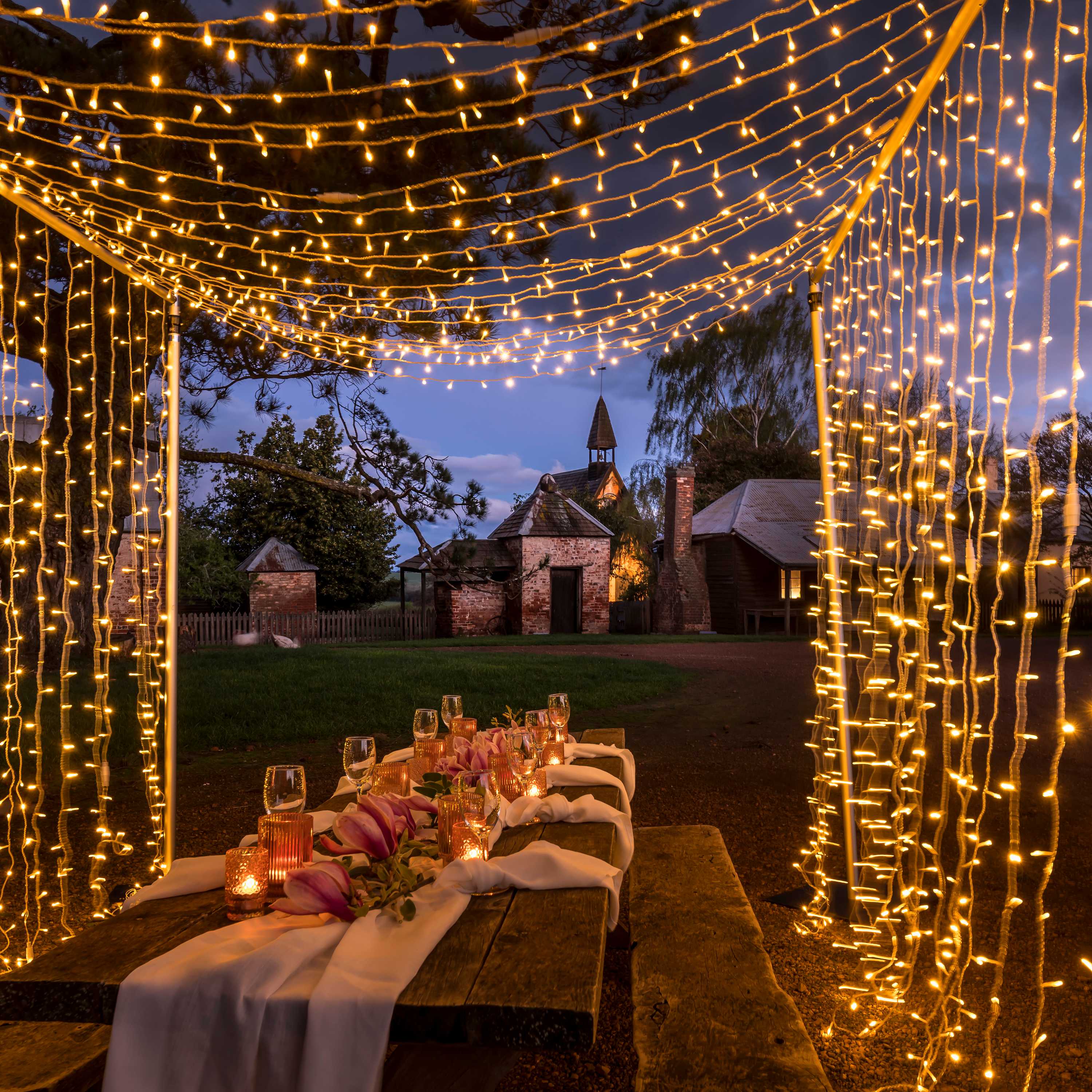 A frame of fairy lights drape over an outdoor table set with wine glasses, flowers and table linen. Farm buildings line the roadway in the background and the sky is starting to darken. Photo: Rob Burnett.