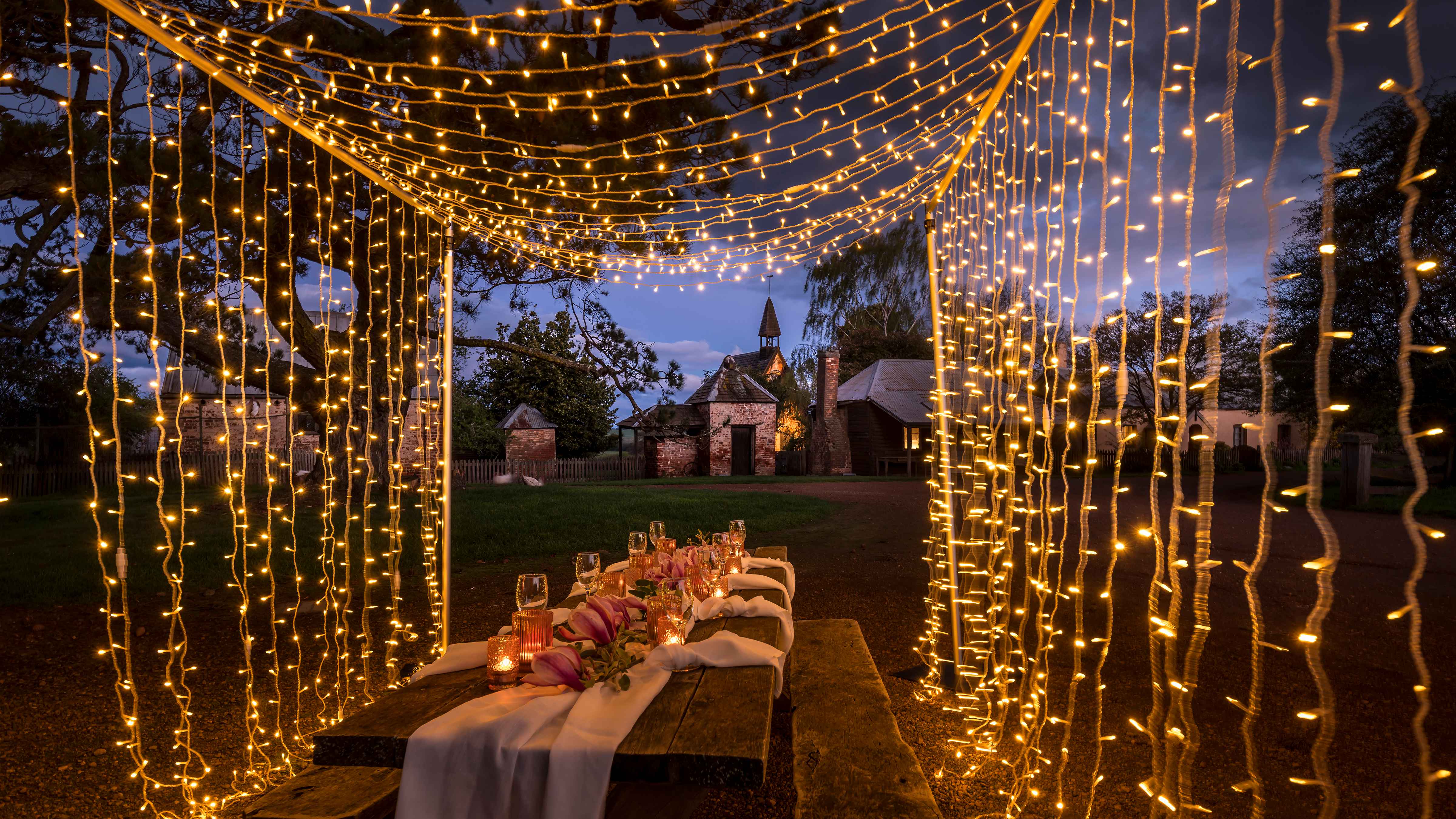 A frame of fairy lights drape over an outdoor table set with wine glasses, flowers and table linen. Farm buildings line the roadway in the background and the sky is starting to darken. Photo: Rob Burnett.