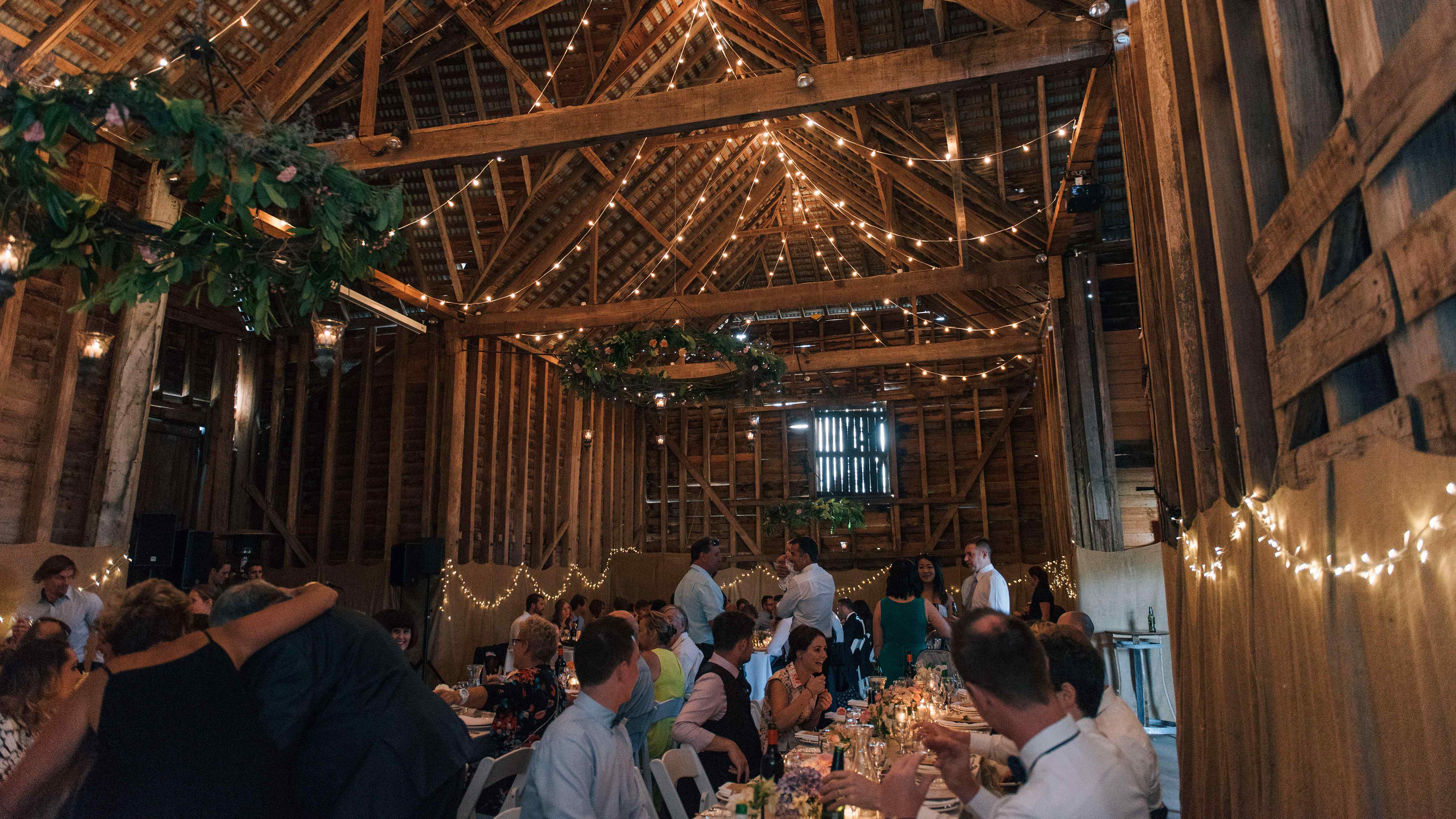 Guests sit at long tables in the Sussex Barn. The ceiling of the barn is draped with festoon lights that show off the grand height of the barn. Three foliage rings hang from the ceiling and more fairy lights are hung around the walls. Photo: Clint & Bethanie Creative.