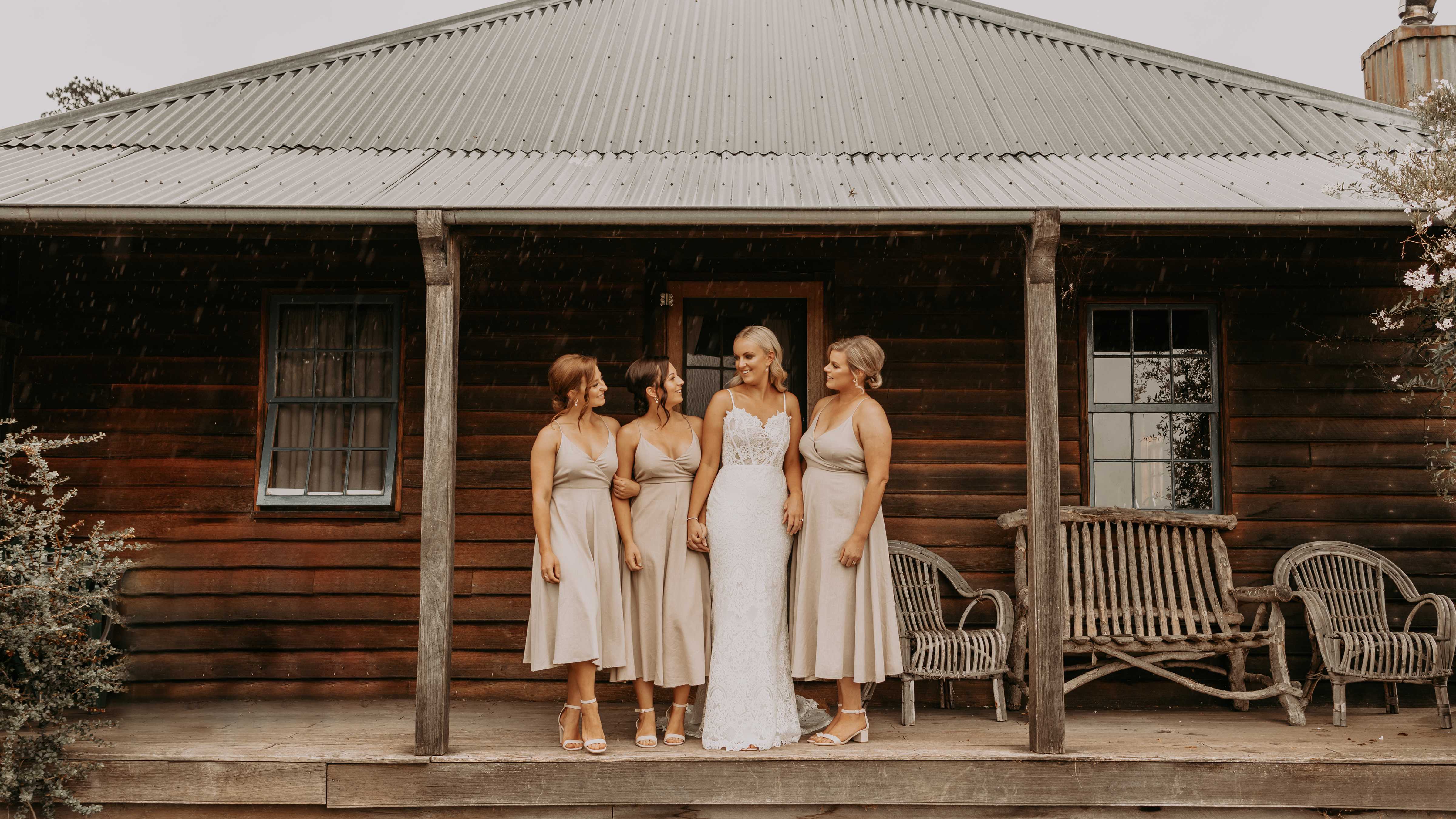 A bride and three bridesmaids in beige dresses stand on the front verandah of Sweetcorn Cottage. The cottage is made from recycled timber with a corrugated iron roof. Rustic timber lounge furniture flank the people. Photo: Tiarne Shaw Photography.