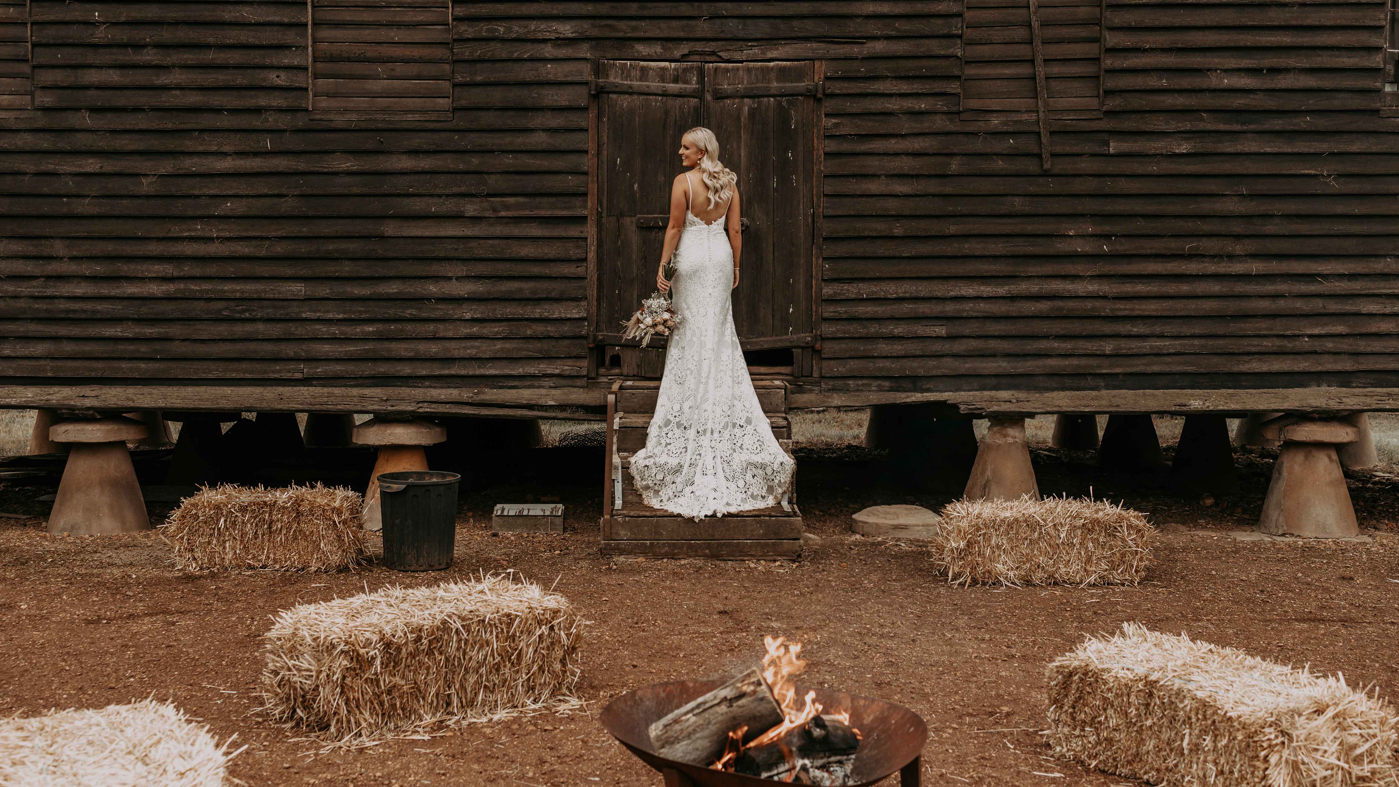 A bride in a wedding dress stands on the steps of the timber Pillar Granary. Hay bales are placed on the gravel below as well as a lit fire pit. Photo: Tiarne Shaw Photography.