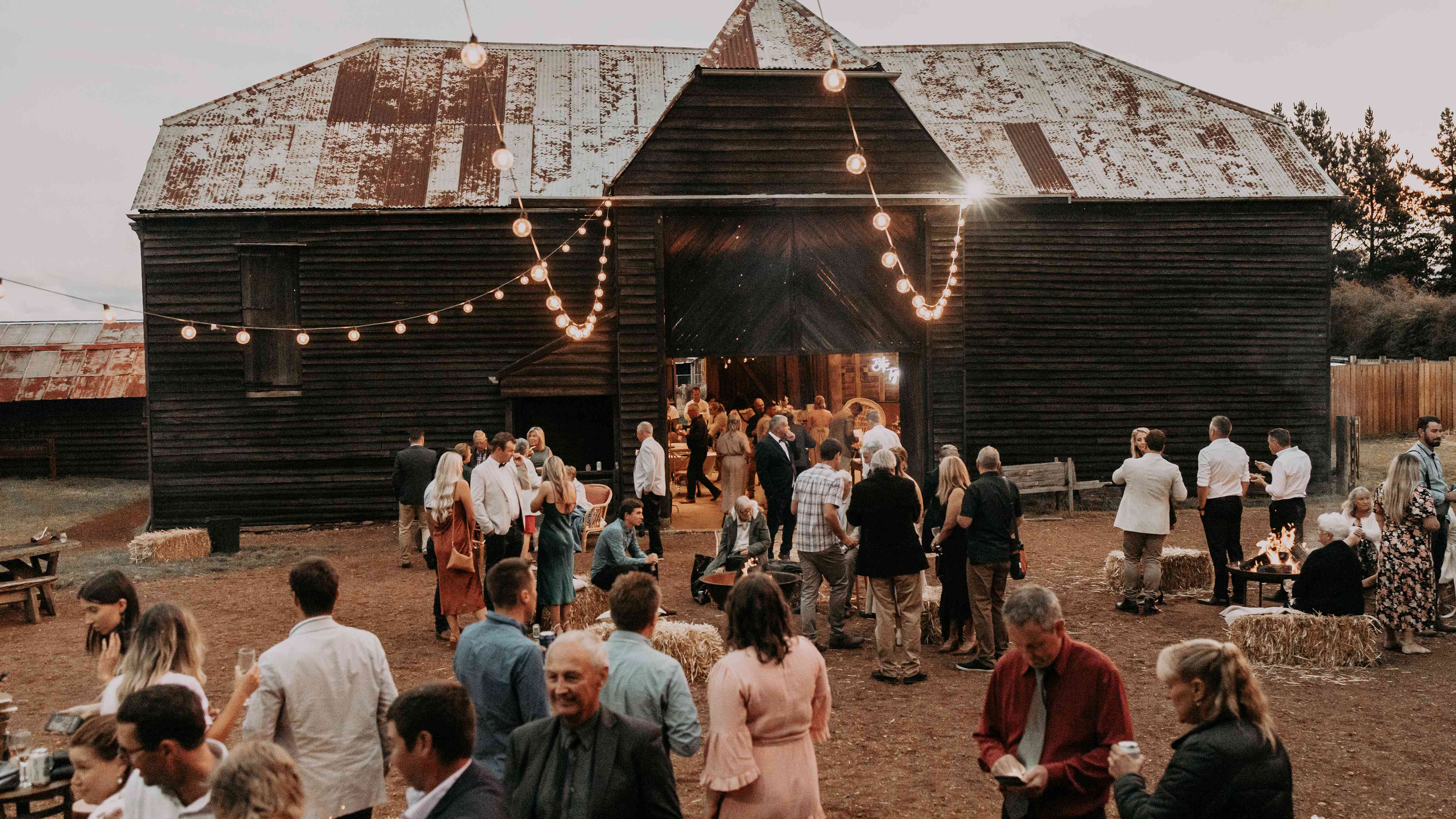 The Sussex timber barn and quadrangle is set for an outdoor wedding reception. Hay bales, fire pits and festoon lights decorate the area along with timber outdoor tables. Guests are mingling outside and inside the Barn. Photo: Tiarne Shaw Photography.