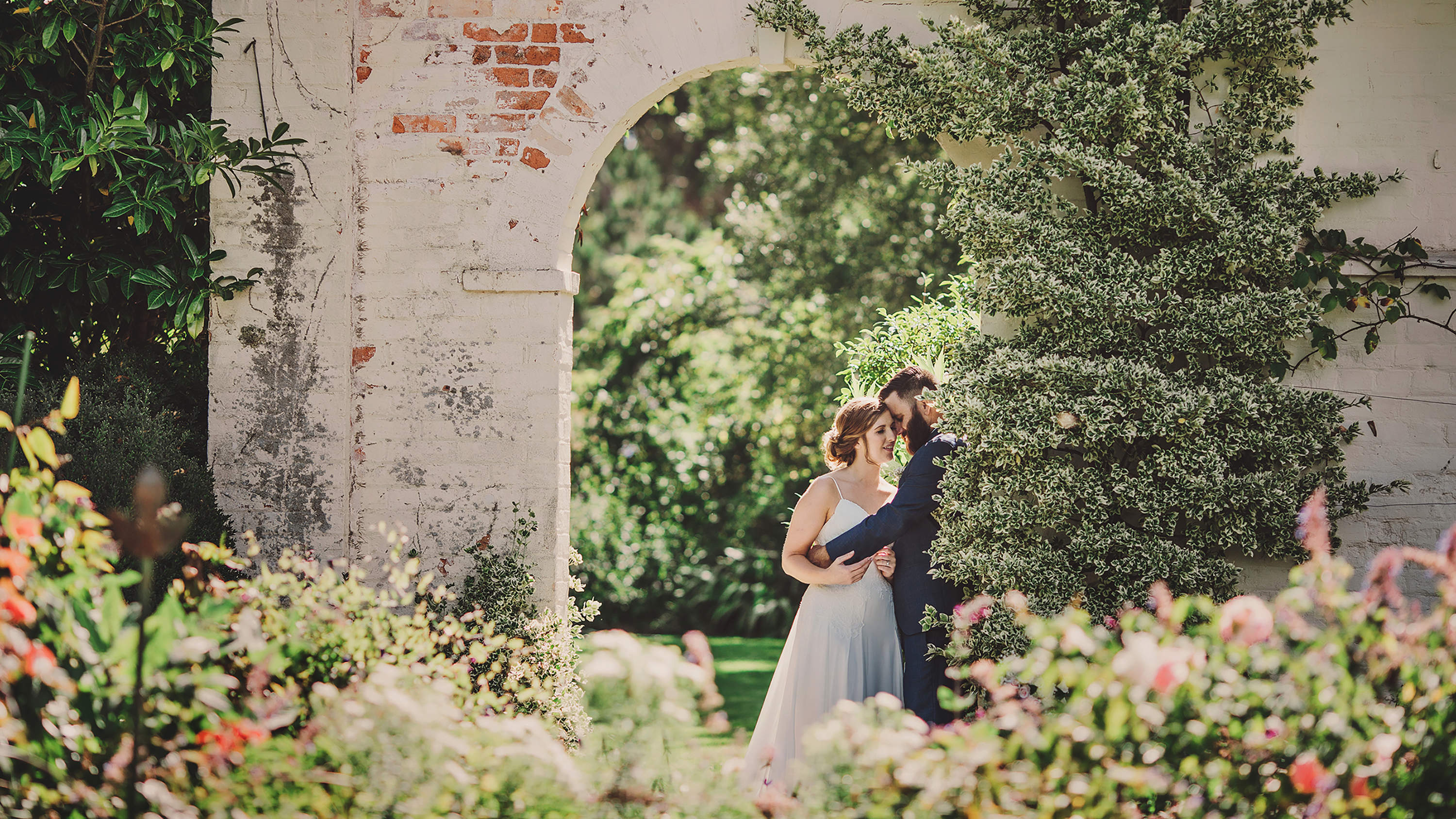 A bride and groom embrace under a white brick archway surrounded by climbing plants and flowers. Photo: Astrid Simone.