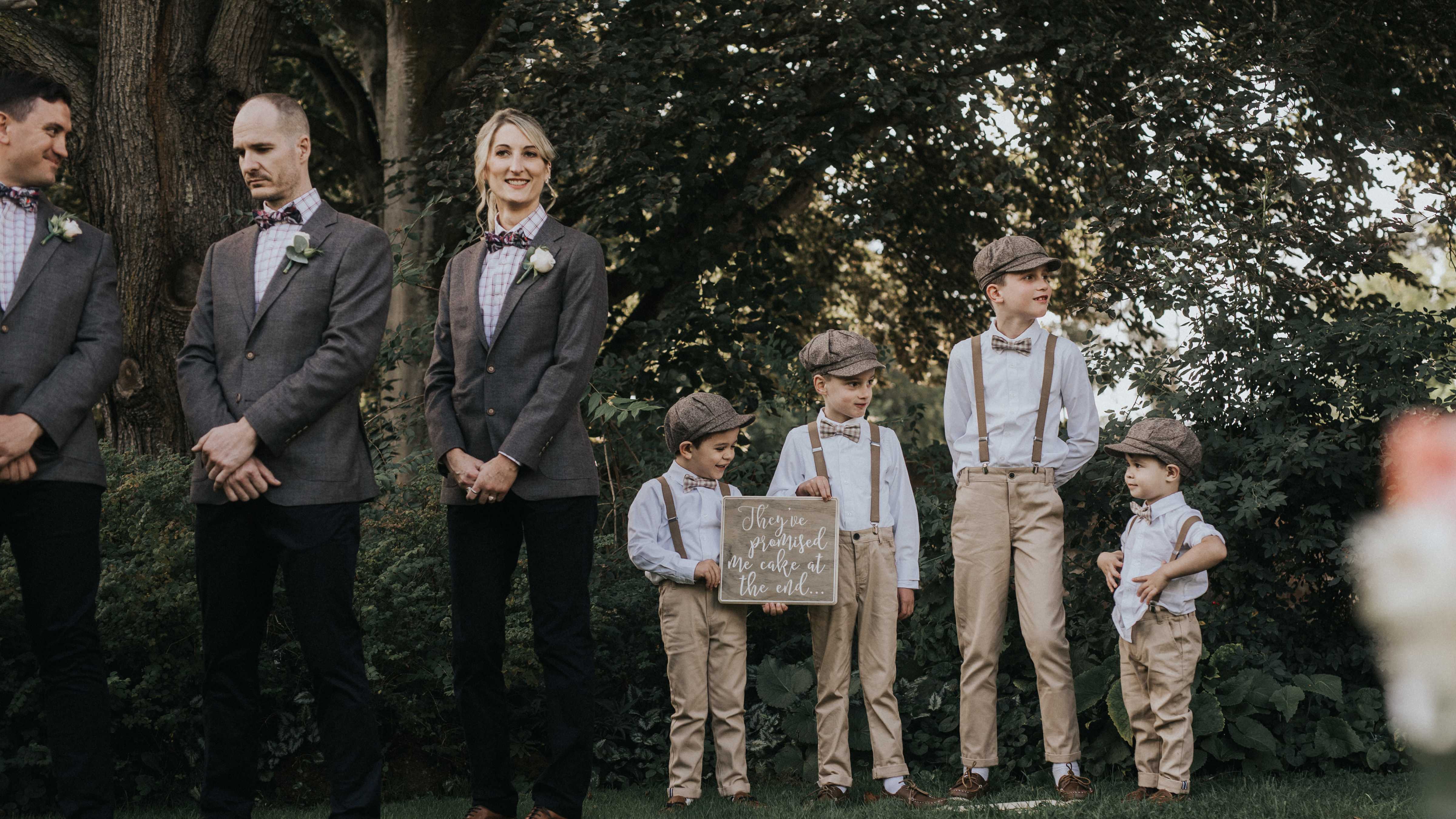 One side of a bridal party including four boys dressed in brown trousers and suspenders stand in front of shrubberies and trees. The boys are holding a wooden sign that says “They&rquo;ve promised me cake at the end”. Photo: Cassie Sullivan.