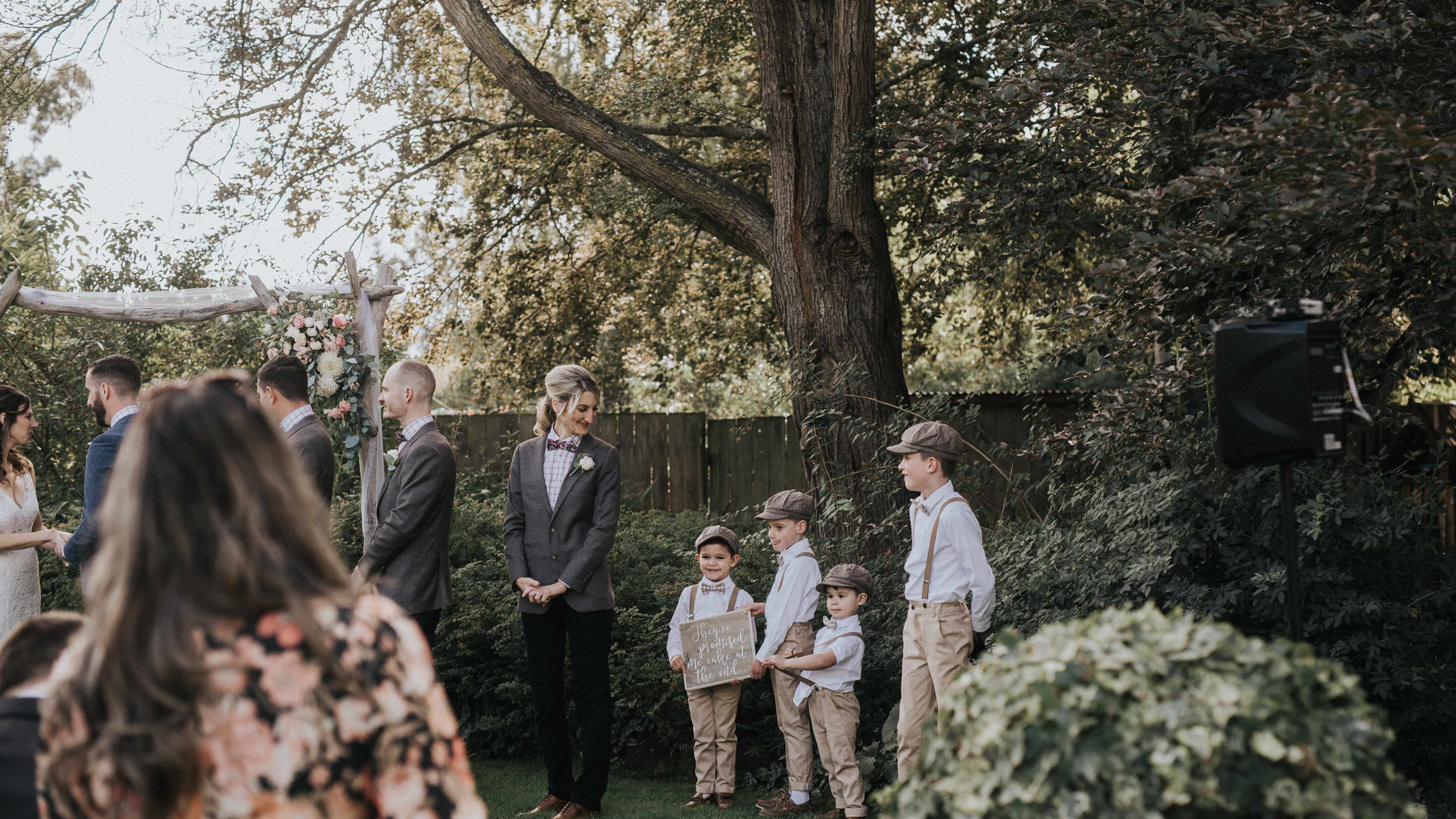 Members of a bridal party stand in front of a wooden arch adorned with flowers. Four page boys are looking cheekily at guests in front of them. Photo: Cassie Sullivan.