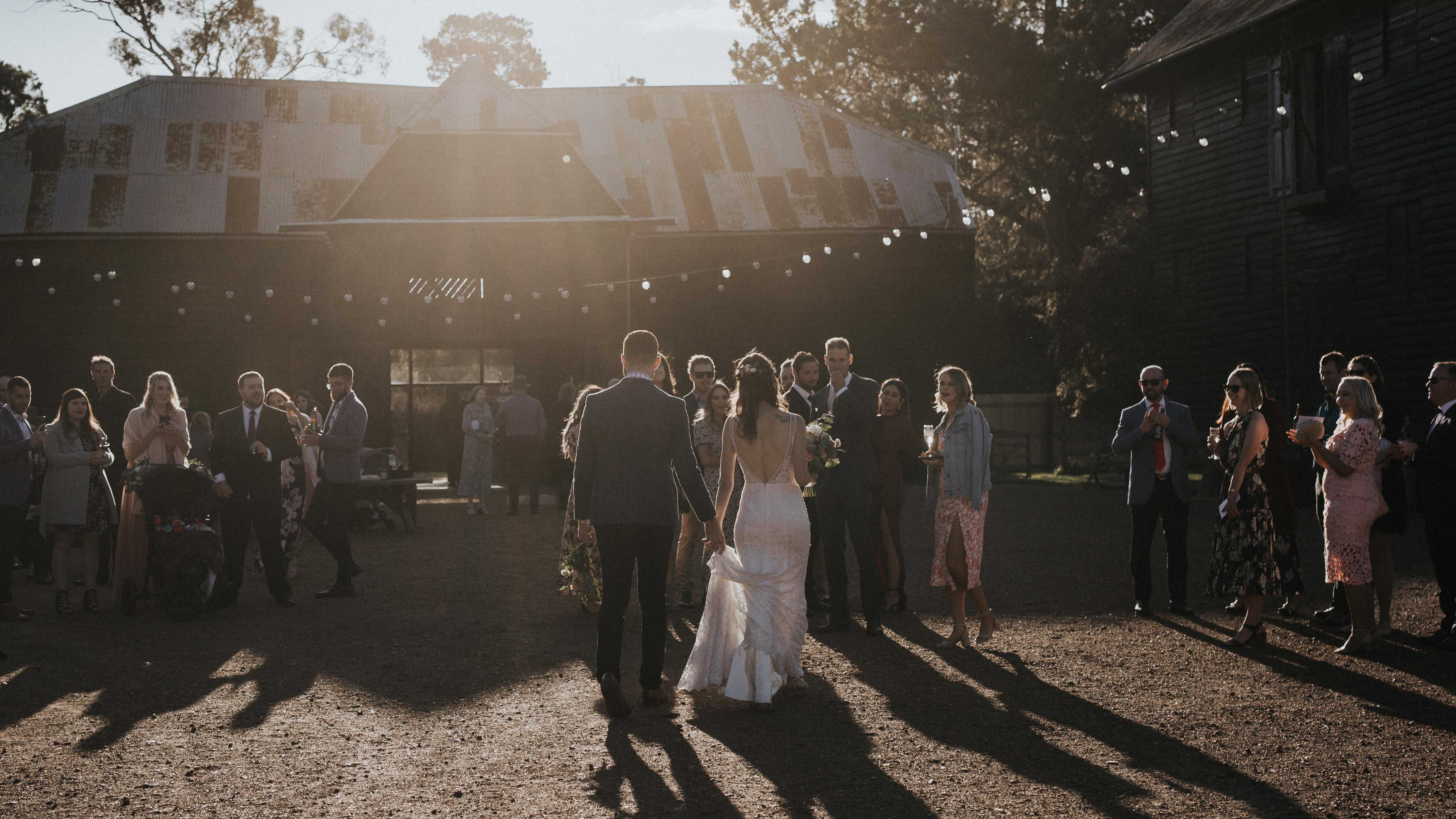 A bride and groom walk towards their guests in the quadrangle. The Sussex Barn sits behind the guests with festoon lights draped in front. Photo: Cassie Sullivan.