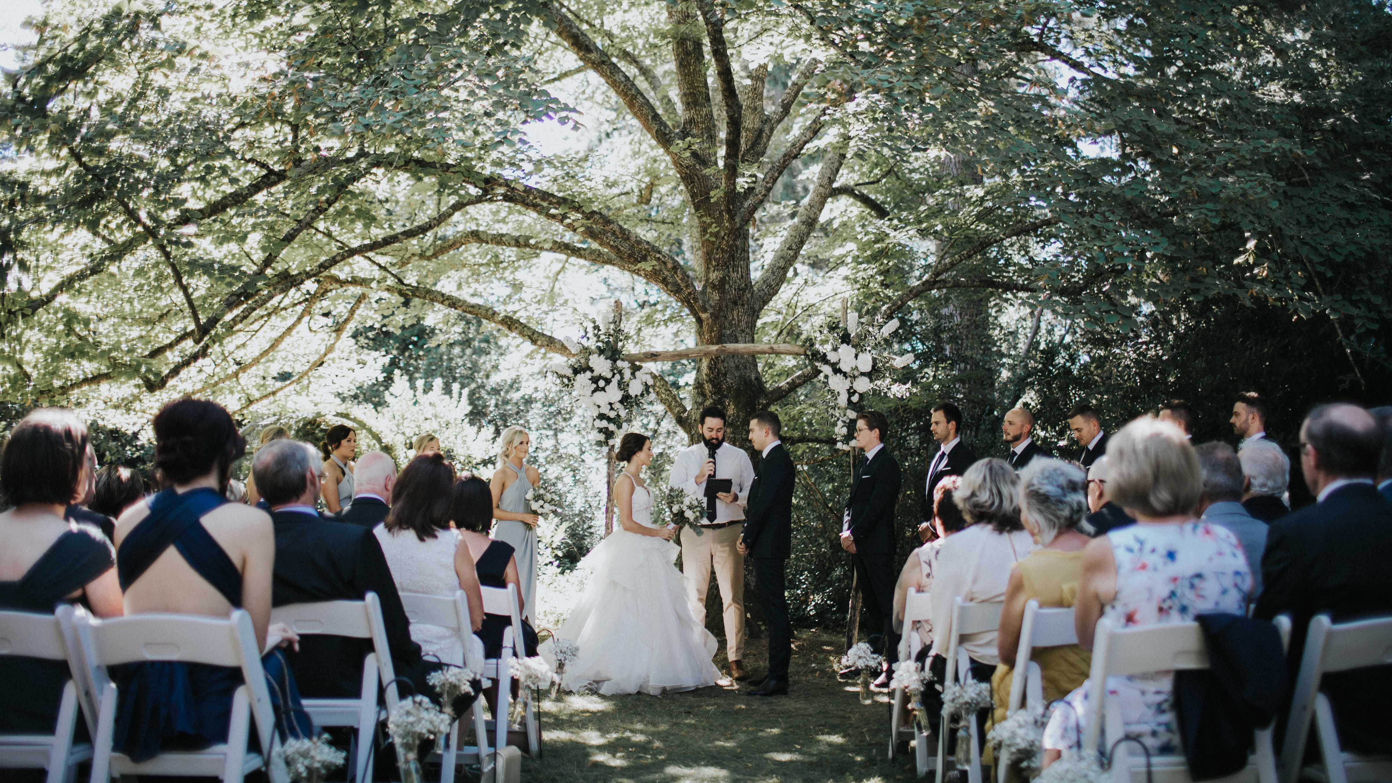 A wedding in full swing beneath the Linden Tree in the Heritage Gardens. A wooden arch adorned with flowers stands behind the bride, groom, and celebrant. Guests are seated on white chairs on either side of the aisle. Photo: Cassie Sullivan.